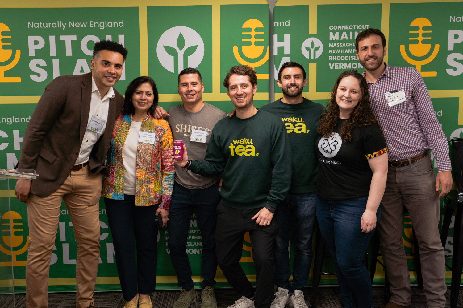 Six people in dark green and other shirts stand close together in front of the Naturally New England Pitch Slam backdrop.