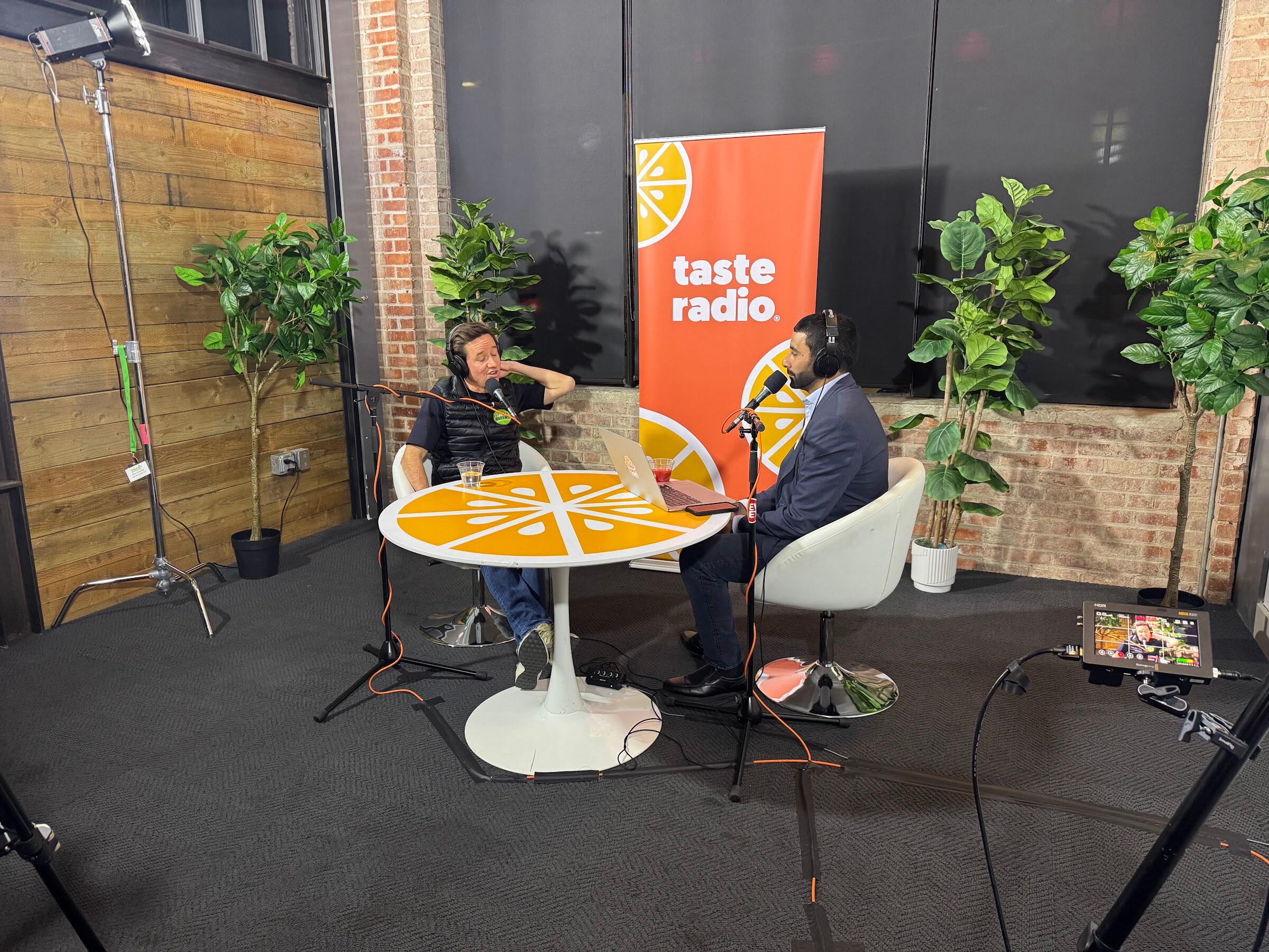 Orange round table, two seated people with headphones, a Taste Radio banner, plants and a brick wall.