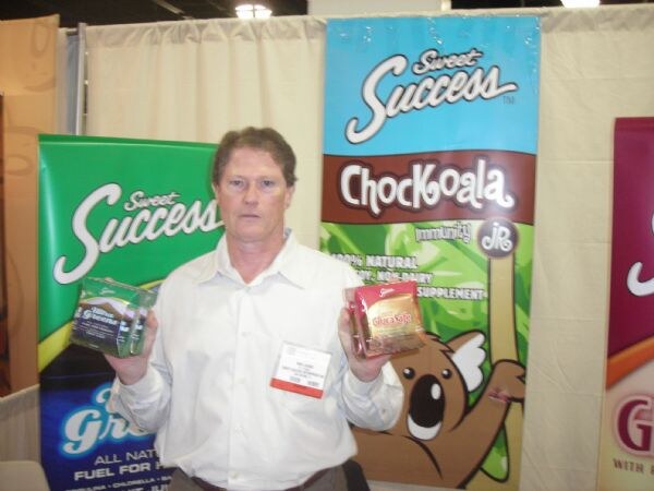 A man in a white shirt holds up two packaged products in front of Sweet Success banners, including a large Chockoala banner with a cartoon koala.