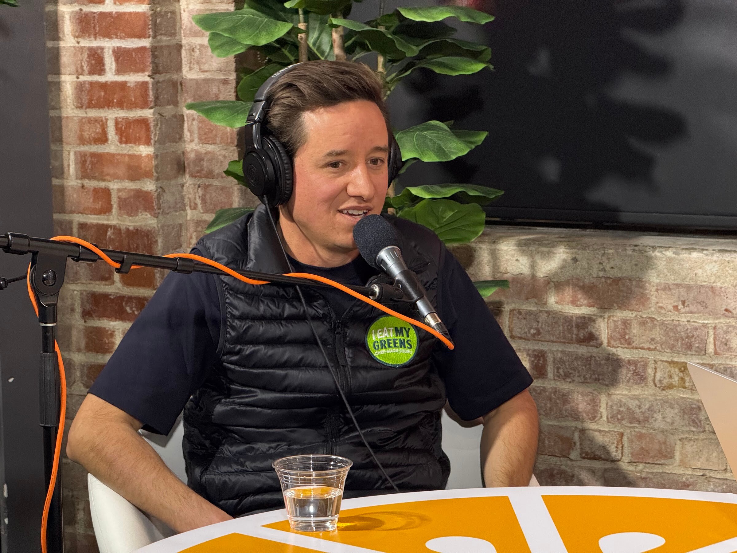 Man in a black puffer vest and headphones speaking into a microphone at an orange table.