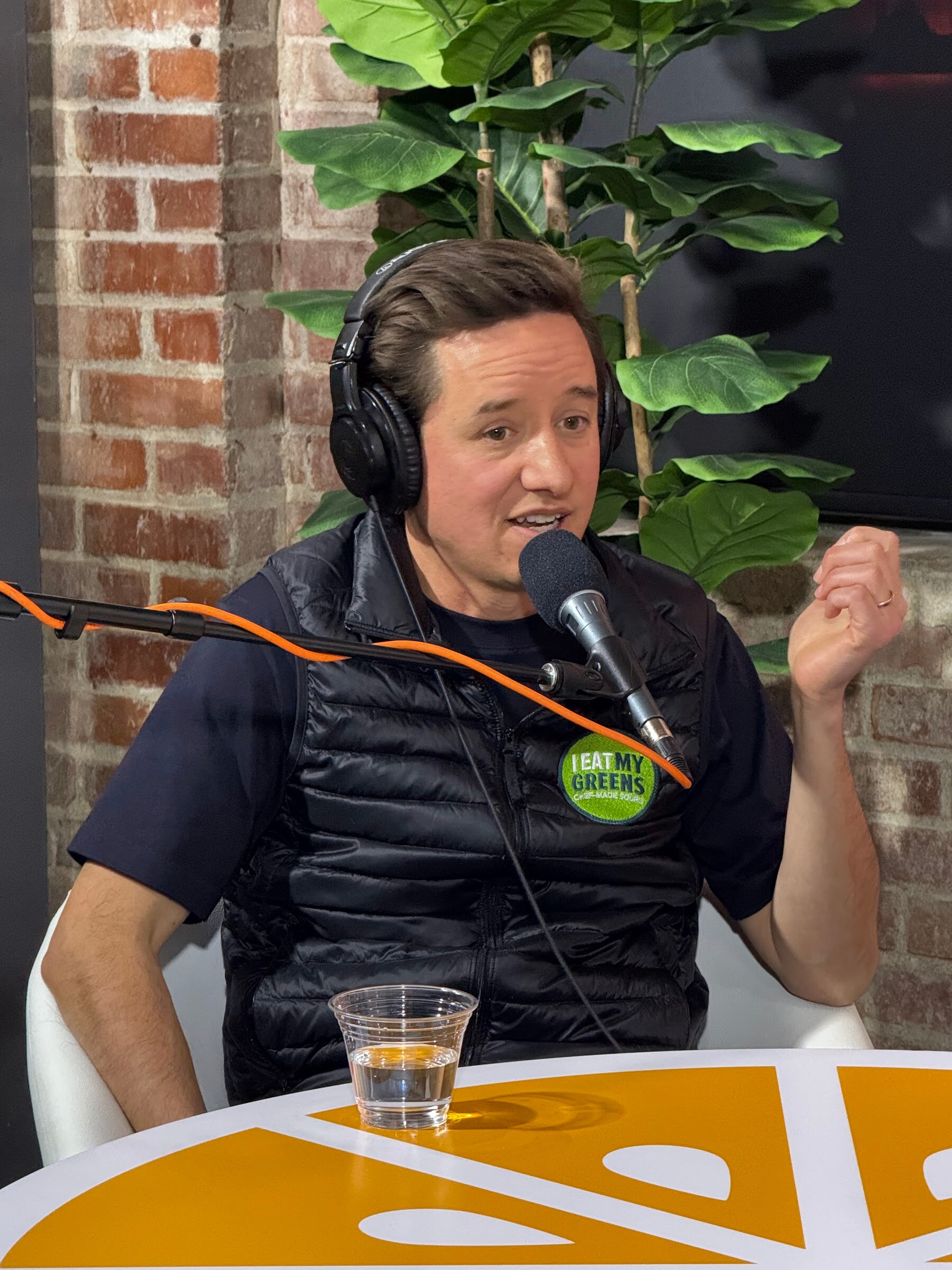 Man in a black vest and headphones with a hand raised, seated at an orange table.