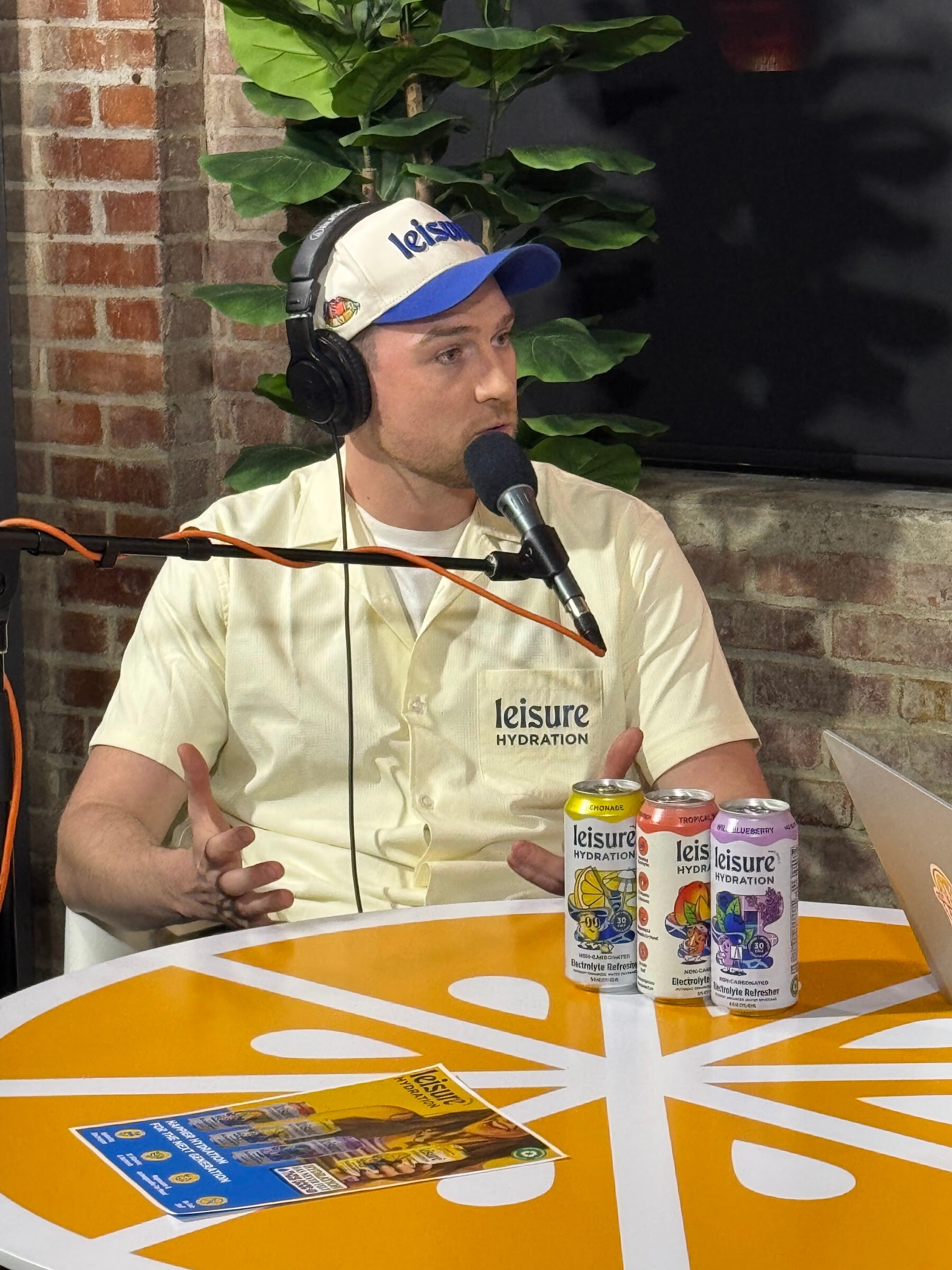 Man in a cream Leisure Hydration shirt and cap speaking, with three cans on an orange table.