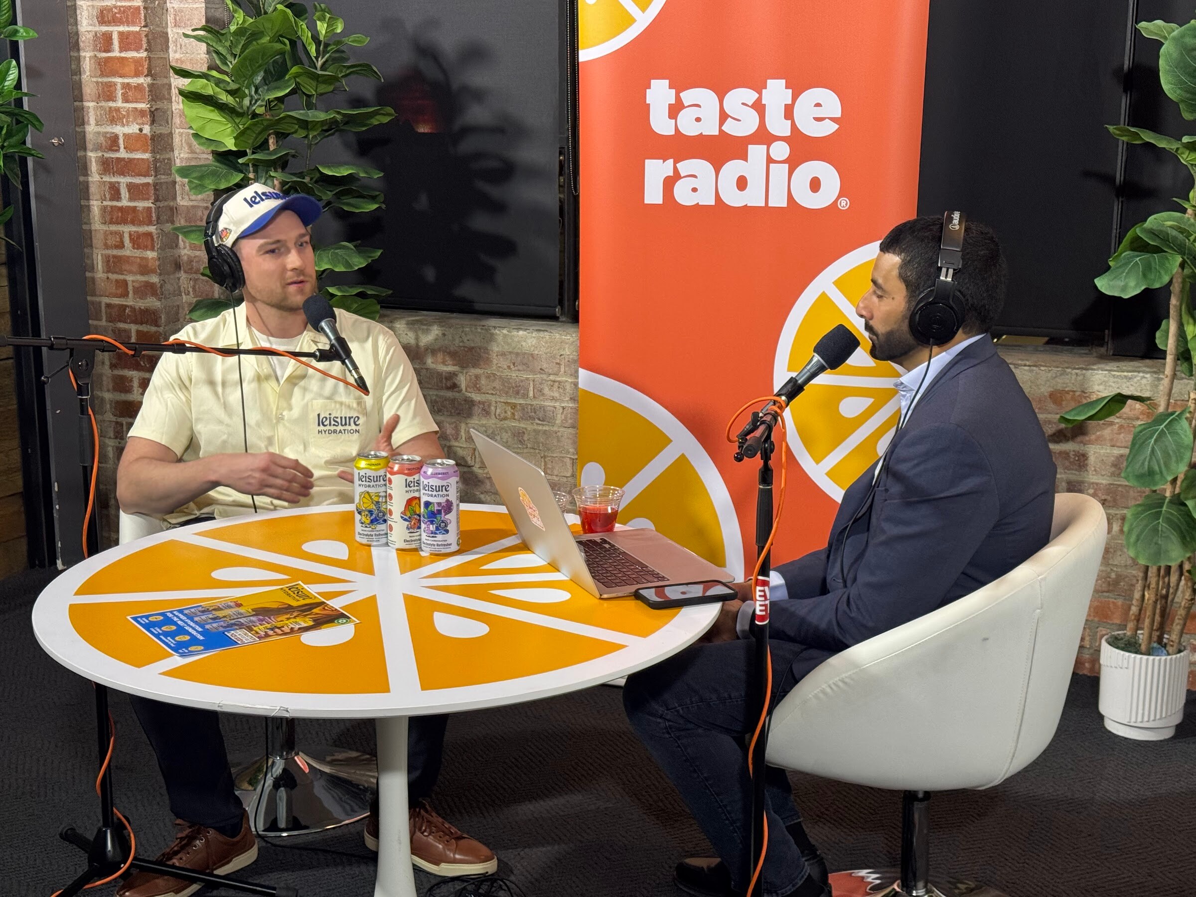 Two men seated at an orange table in front of a Taste Radio banner, with three cans between them.