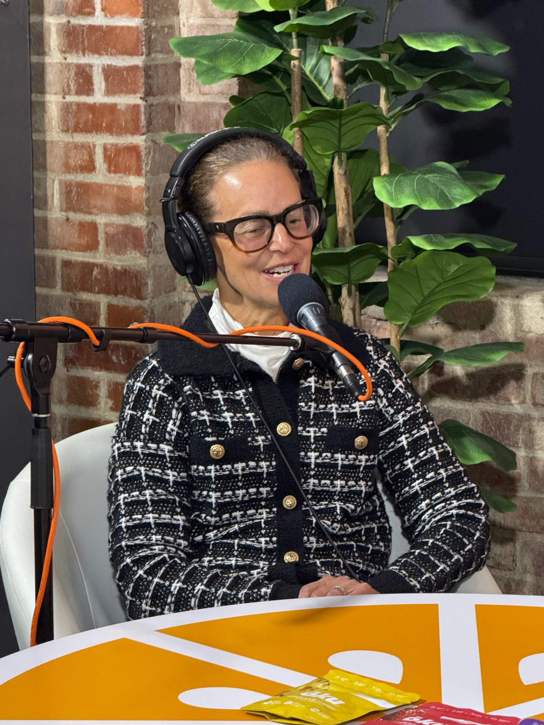 Woman in glasses, headphones and a black and white tweed jacket at an orange table.