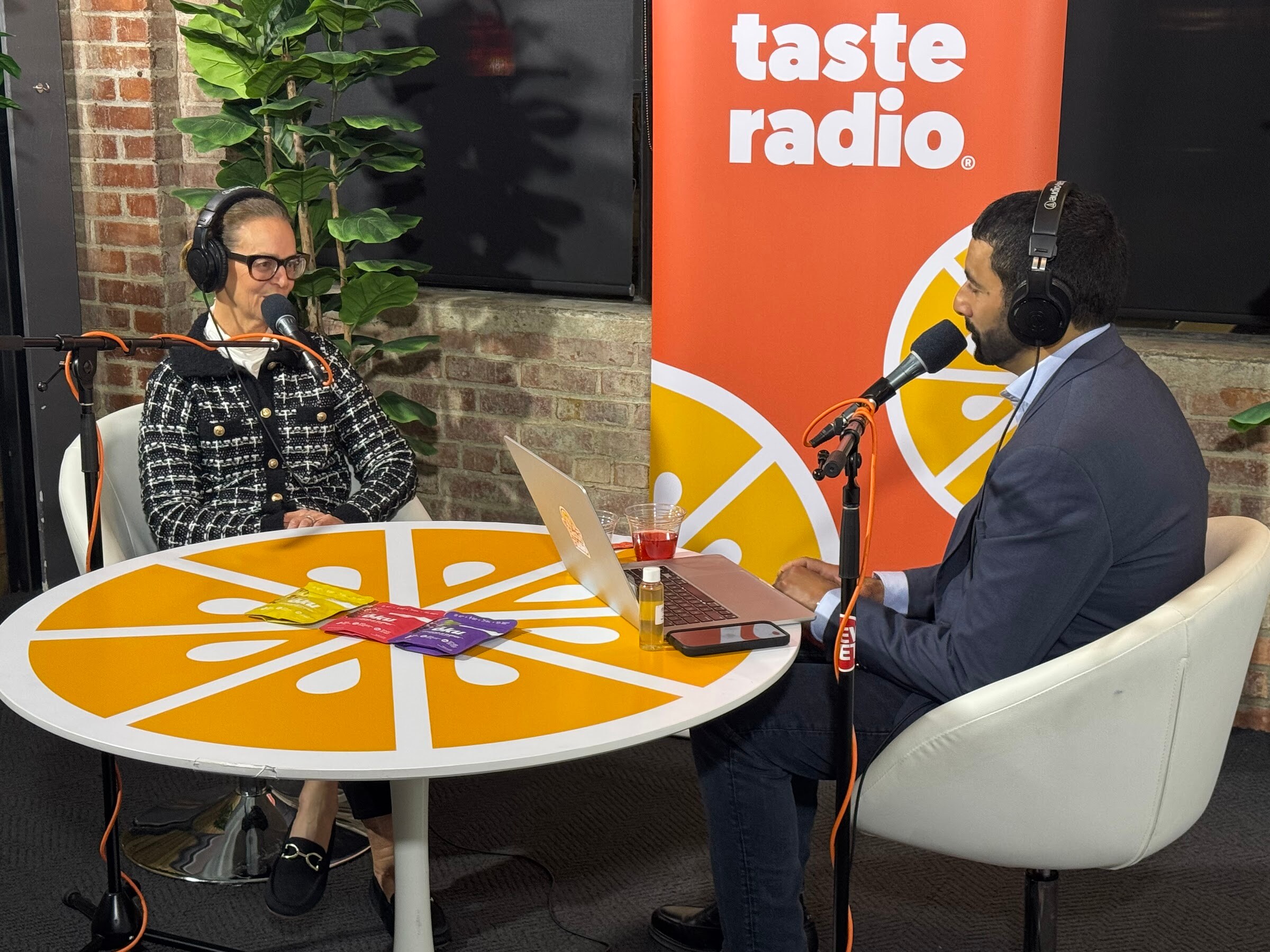Woman with glasses and a man in a navy suit with headphones seated at an orange table in front of the banner.