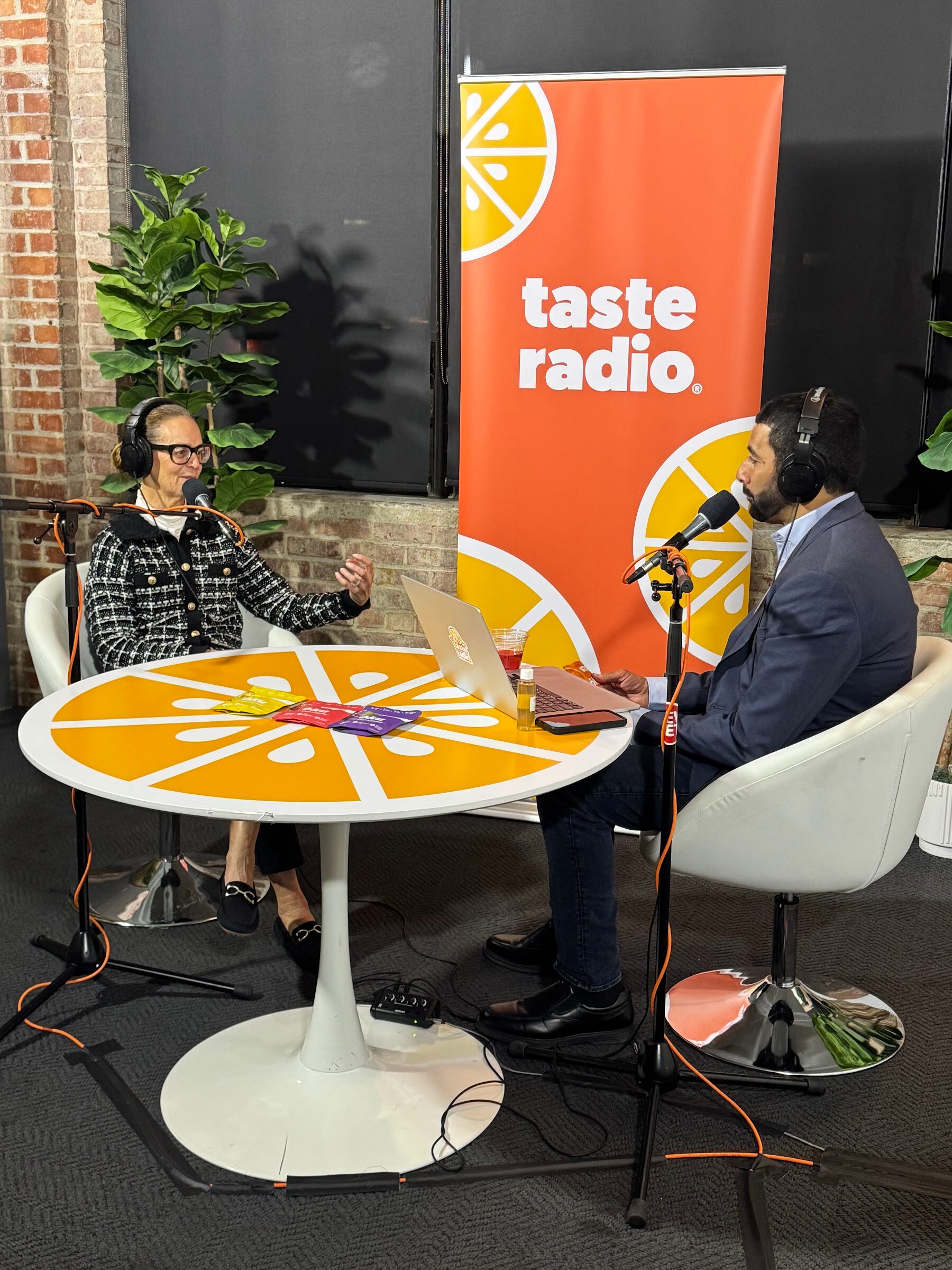 Two people at an orange round table in front of a Taste Radio banner with plants behind.