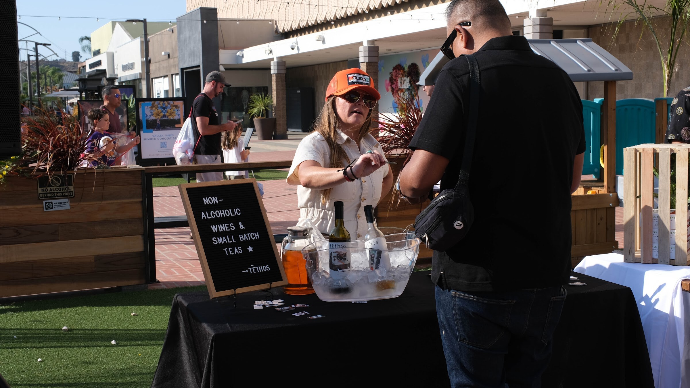A woman in an orange cap stands behind a table with a chalkboard sign reading Non-alcoholic wines, small batch teas, talking with a man in a black shirt.