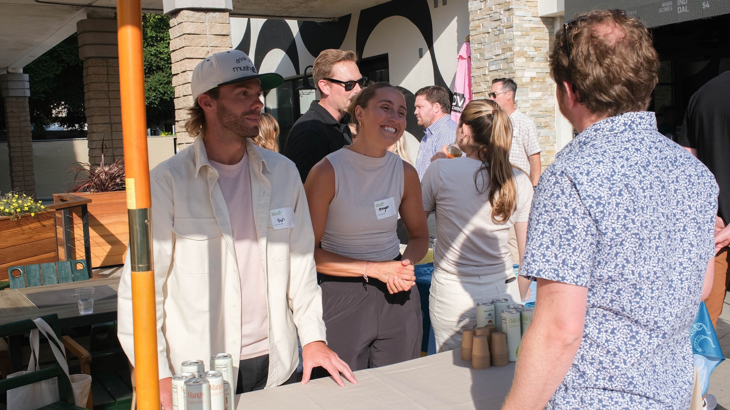 A group of smiling guests with name tags stand at a table of bottles under a covered walkway.