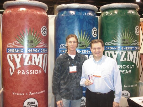 Two men stand in front of large inflatable Blue Agave Syzmo organic energy drink cans in Passion and Prickly Pear flavors. One man holds a small can.
