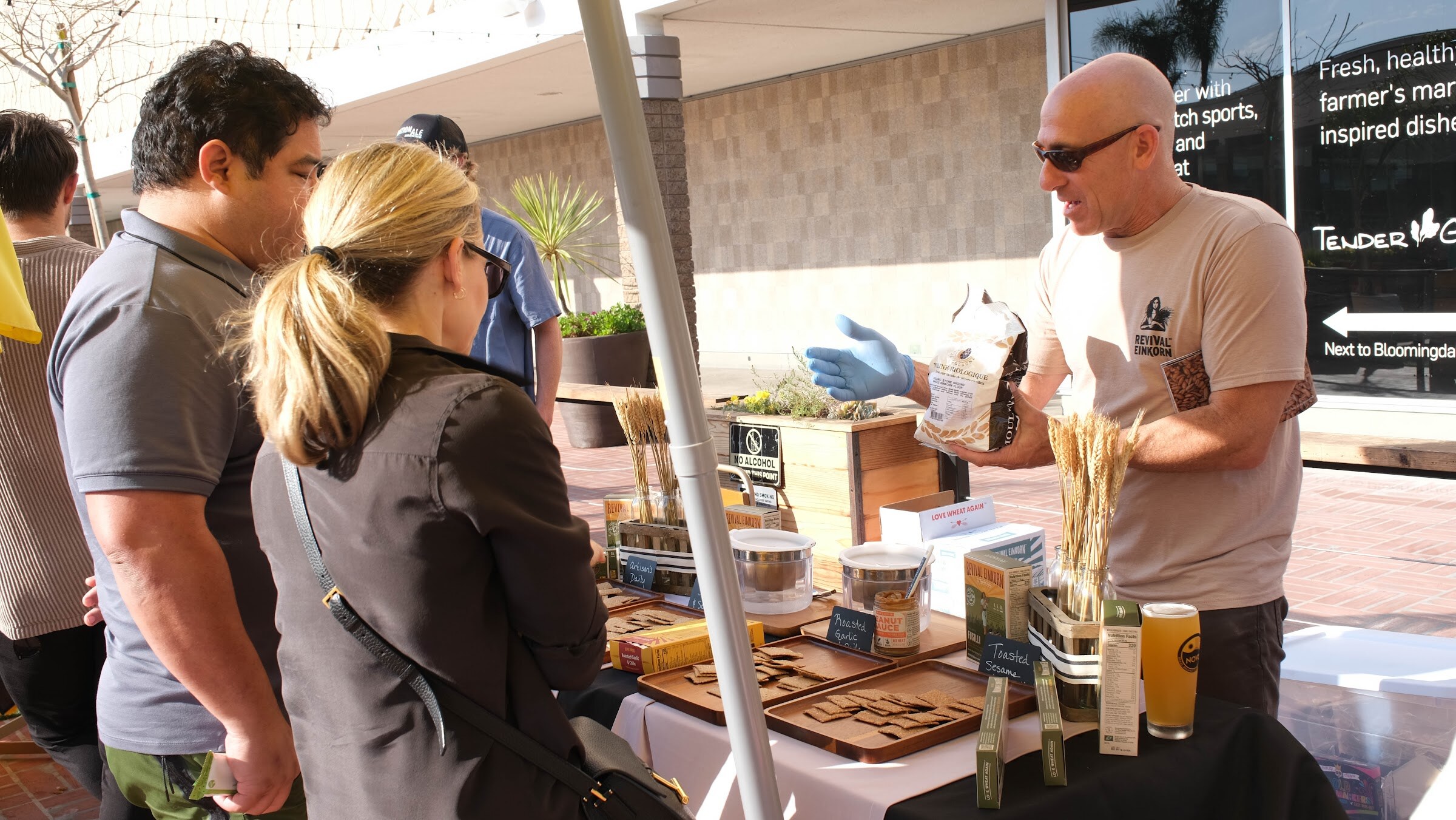 A man in a tan T-shirt and blue gloves holds up a package at a table of jars and wooden trays while two guests look on.