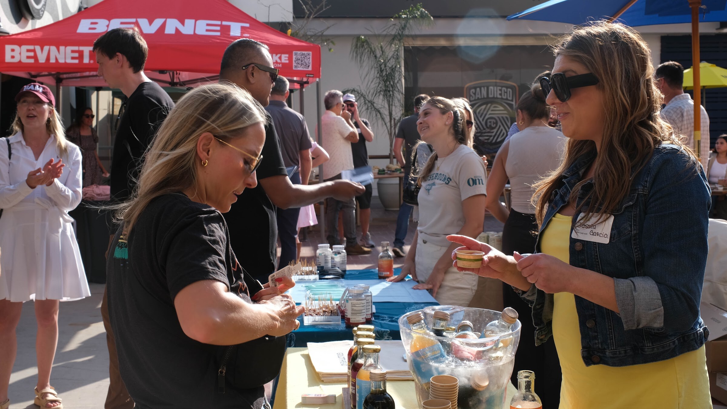 Two women in sunglasses sample products at a table, with a red BevNET canopy and other guests behind them.