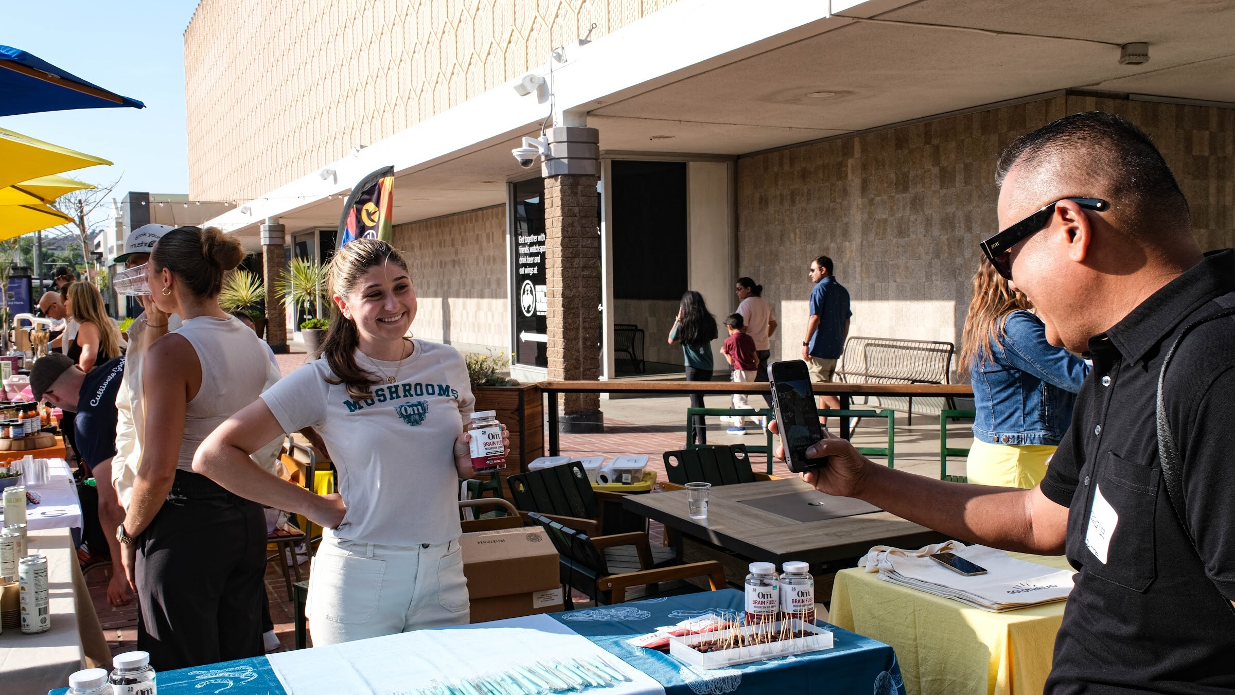 A smiling woman in a white Mushrooms T-shirt stands at a blue-clothed table while a man in sunglasses photographs it with his phone.