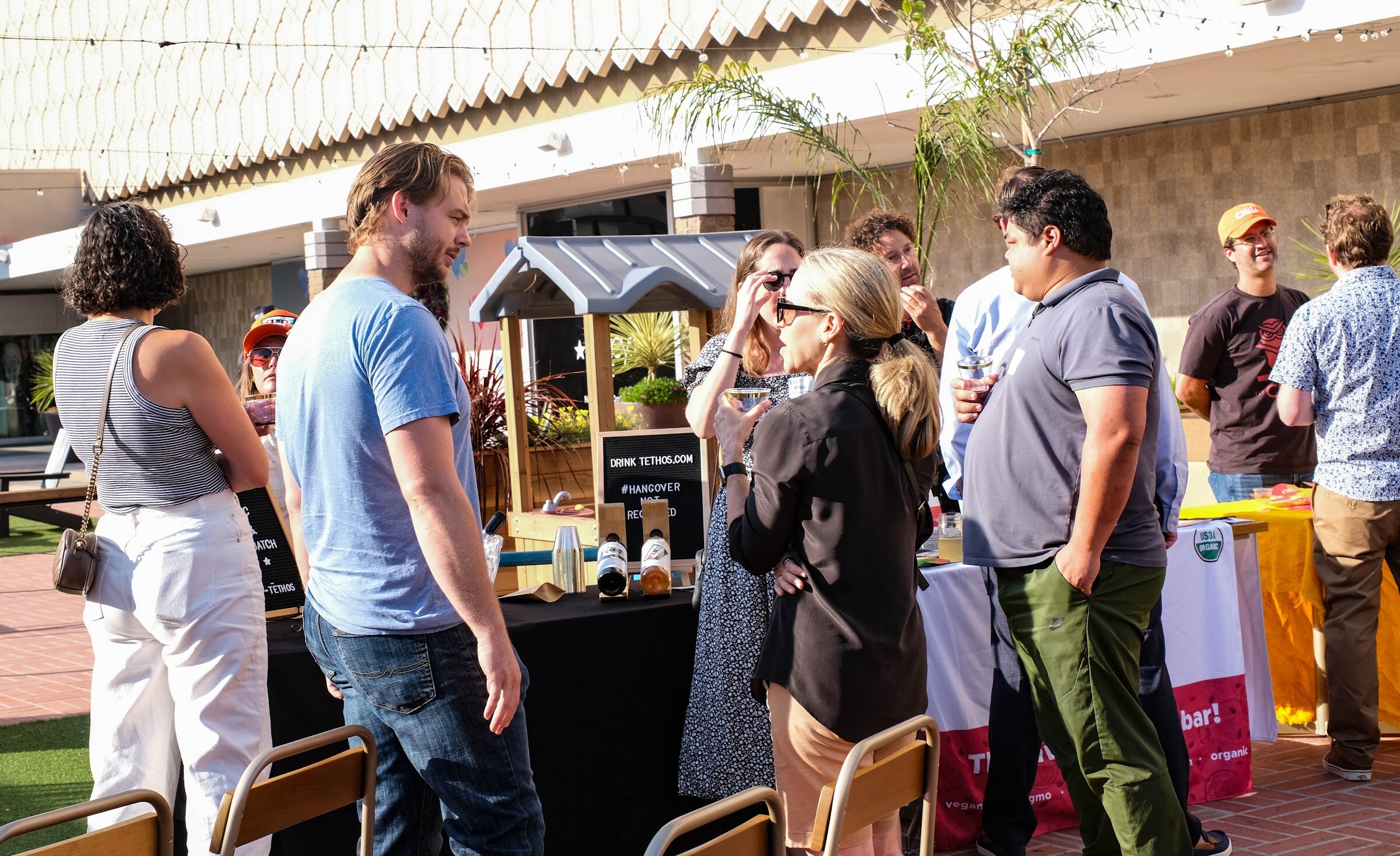 A crowd of attendees talking in a sunny courtyard beside a wooden playhouse and several vendor tables.