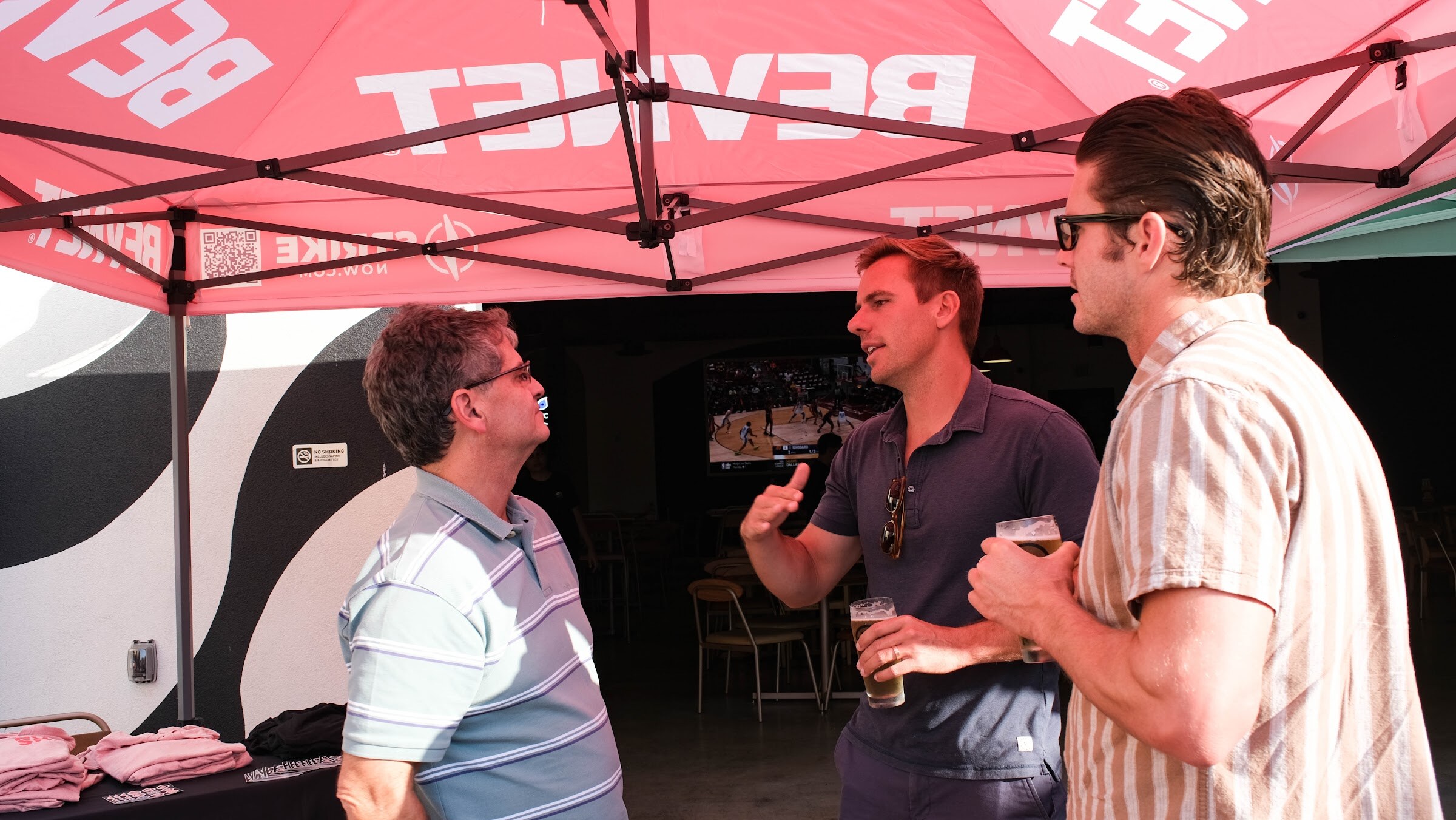 Three men talk with drinks in hand under a pink BevNET canopy, with a patterned wall behind them.