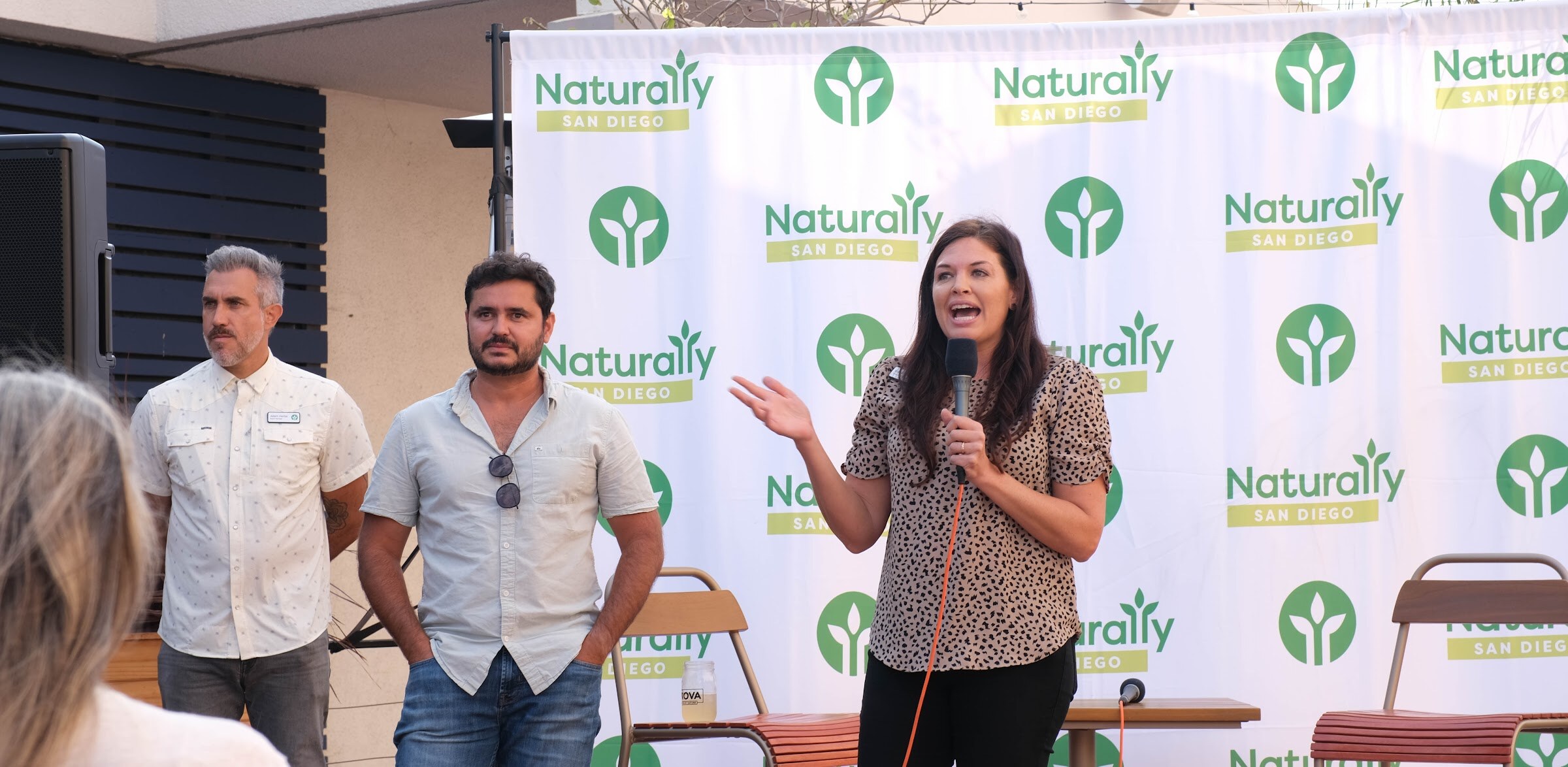 A woman holding a microphone speaks beside two men in front of a white Naturally San Diego step-and-repeat banner.