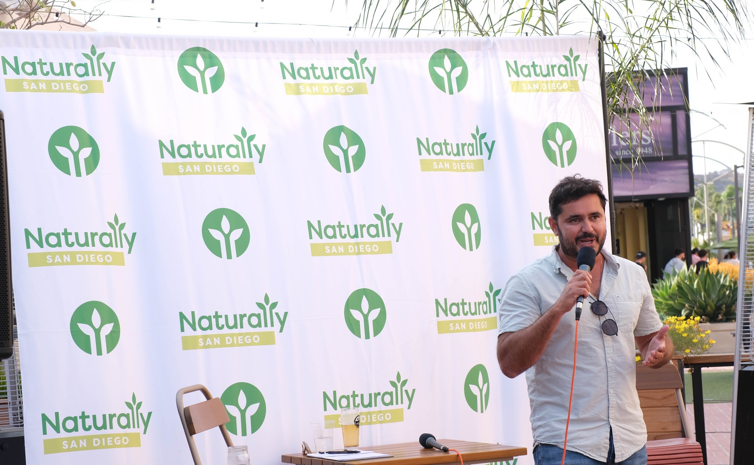 A man in a light shirt and sunglasses holds a microphone and gestures in front of a Naturally San Diego banner.