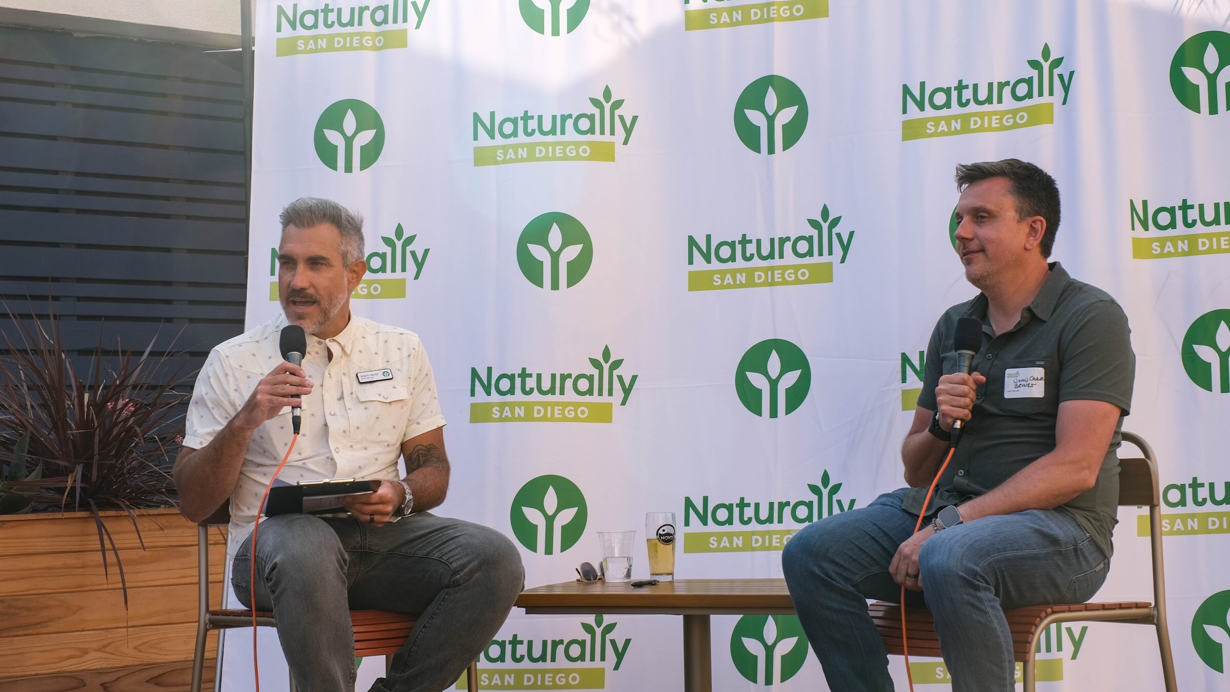 Two men sit on stools holding microphones in front of the white Naturally San Diego banner, one holding a notepad.