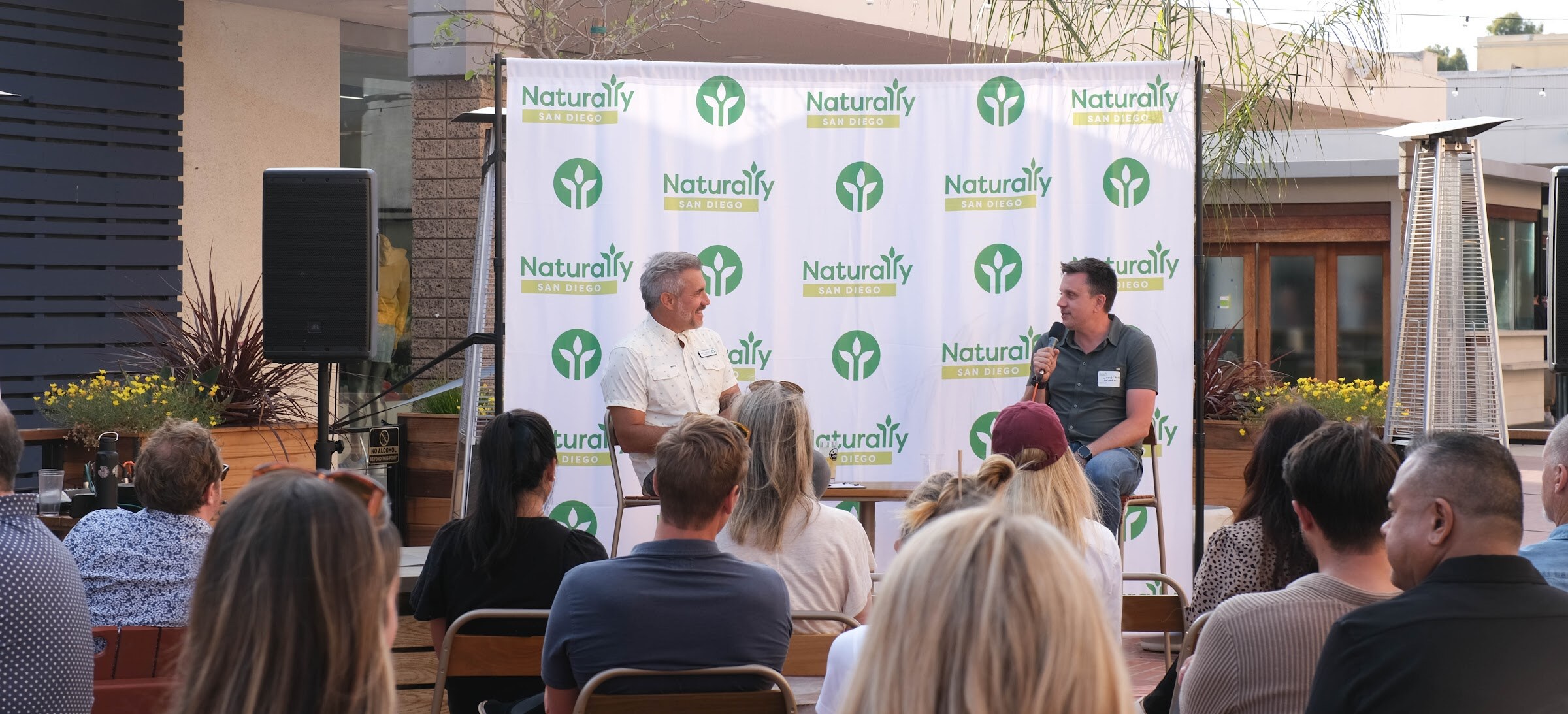 A wide view of two men on stools in front of the Naturally San Diego banner with a seated audience in the foreground and a patio heater beside the stage.
