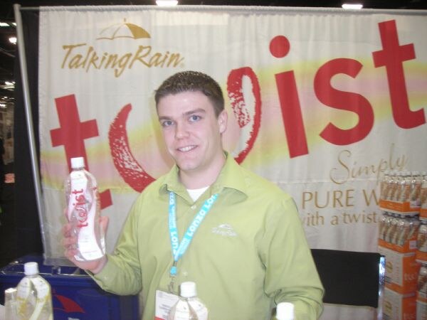 A smiling man in a green shirt holds a bottle of Twist water in front of a Talking Rain Twist banner. More bottles and cases are visible nearby.