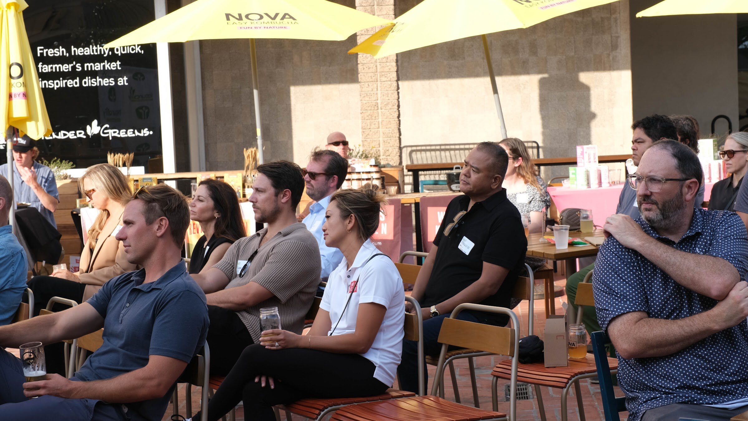 A crowd of attendees seated and standing in a sunny courtyard, watching the stage.