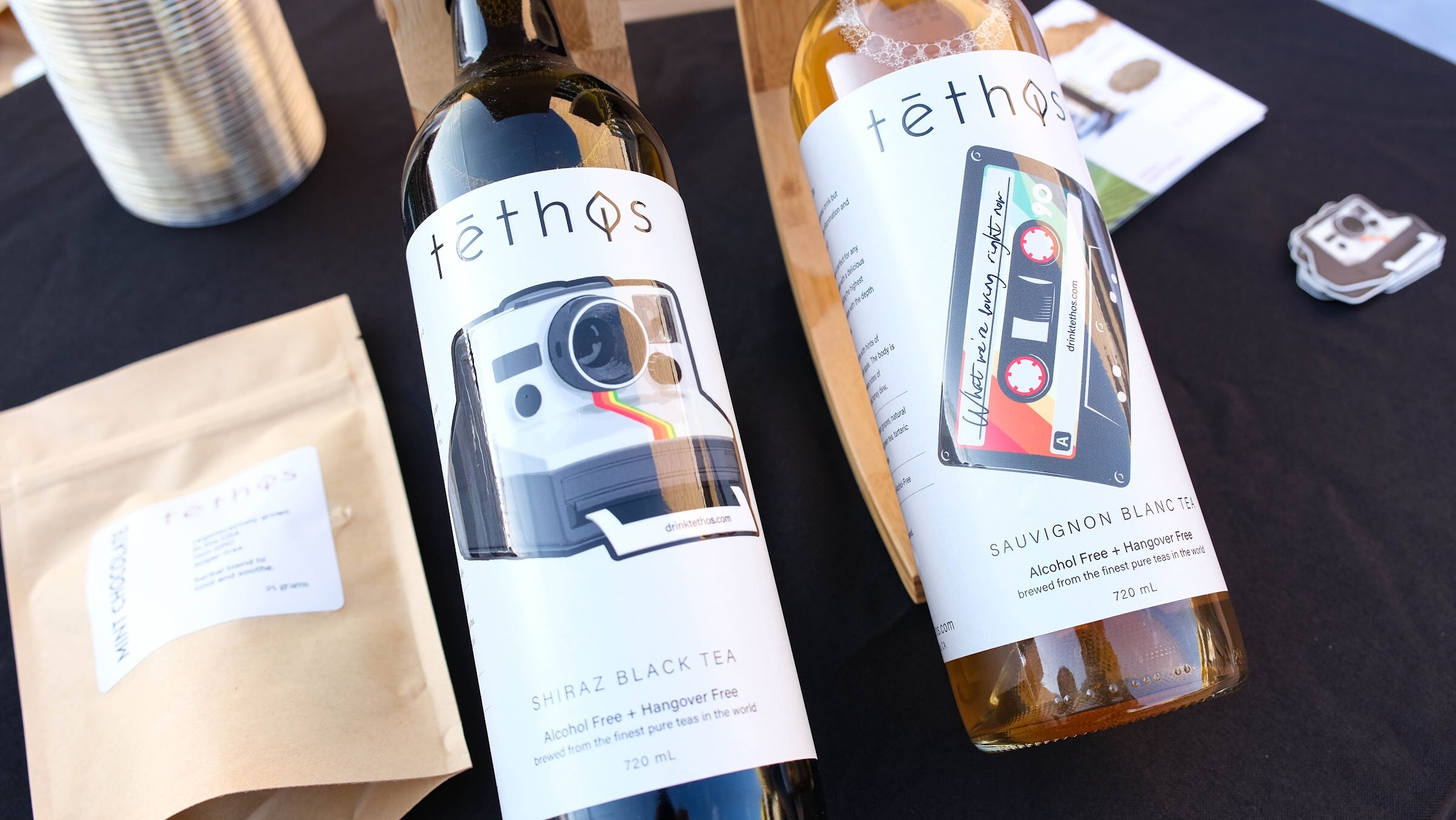 Two bottles labeled tethos, a Shiraz black tea and a Sauvignon blanc, with a tea pouch on a black tablecloth.