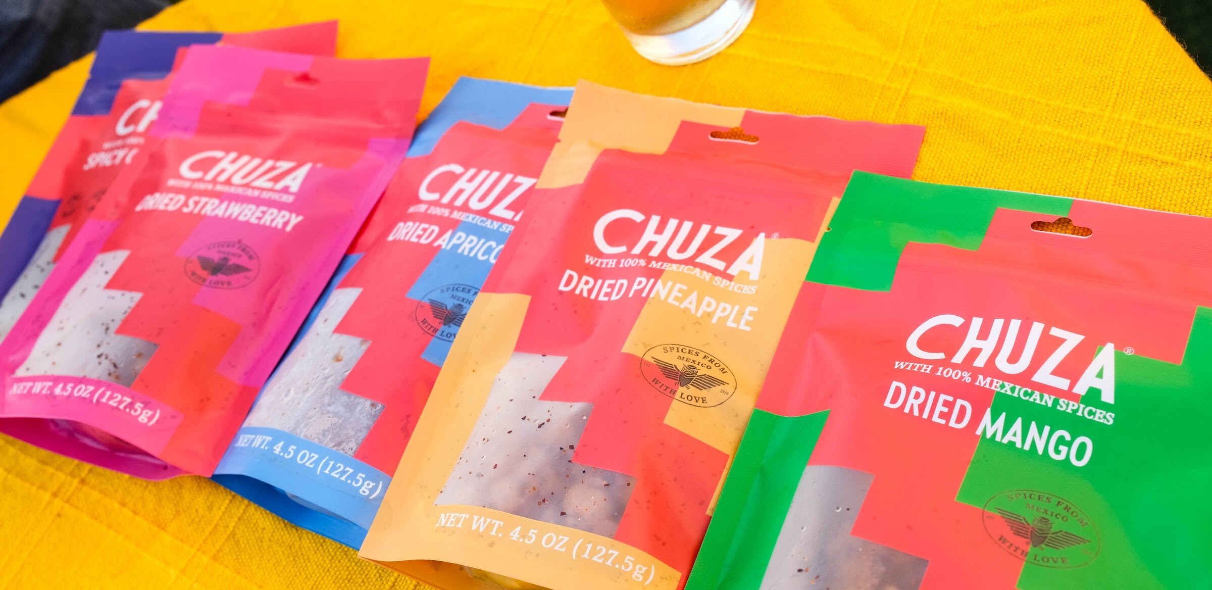 Brightly colored bags of Chuza dried fruit in pink, orange and green fanned out on a yellow tablecloth.
