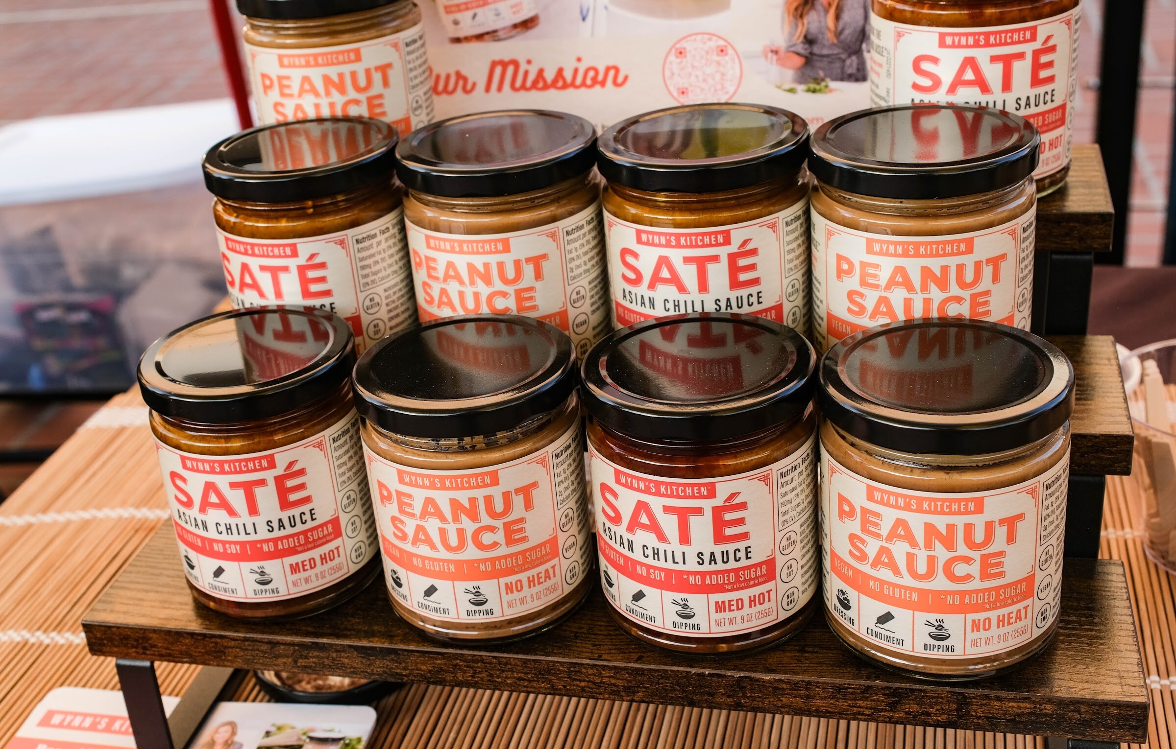 Eight jars of Saté Asian chili sauce and peanut sauce stacked on a wooden display riser.