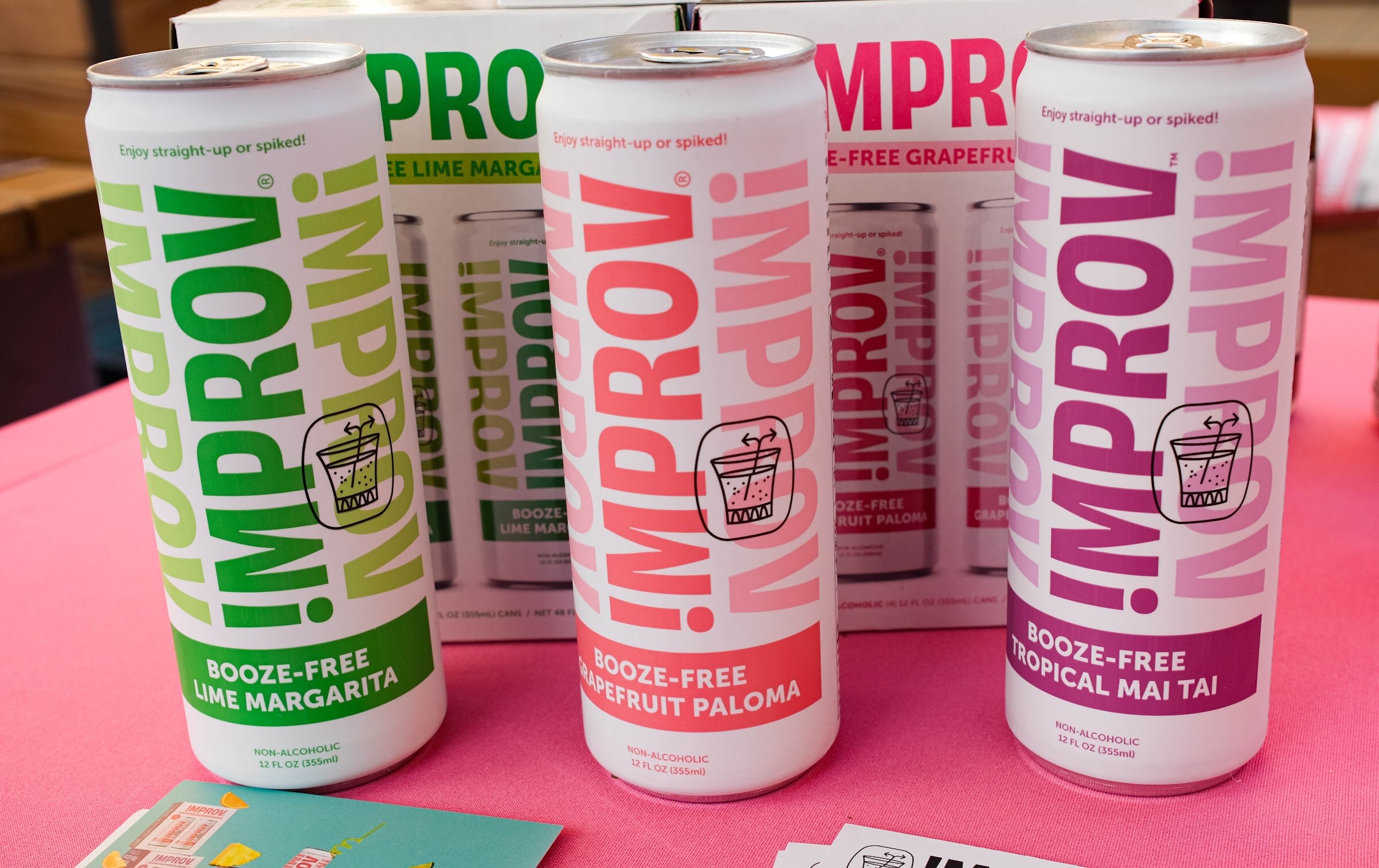 Three Improv cans in lime margarita, grapefruit paloma and tropical mai tai flavors on a pink tablecloth.