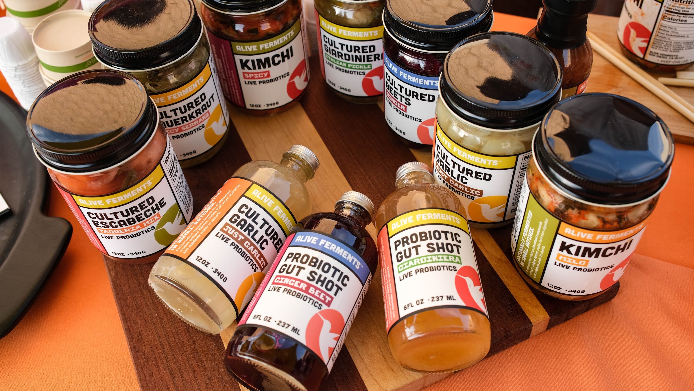 Bottles of cultured chili sauce and probiotic shots and jars of kimchi arranged on a wooden board over an orange cloth.