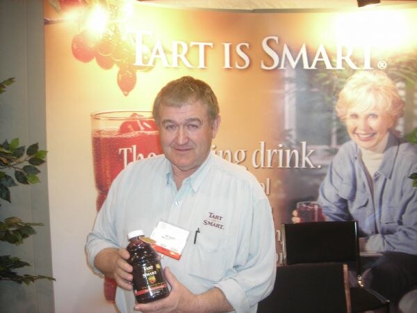 A man in a light blue Tart is Smart shirt holds a bottle of dark juice in front of a Tart is Smart backdrop showing a smiling woman with a glass.