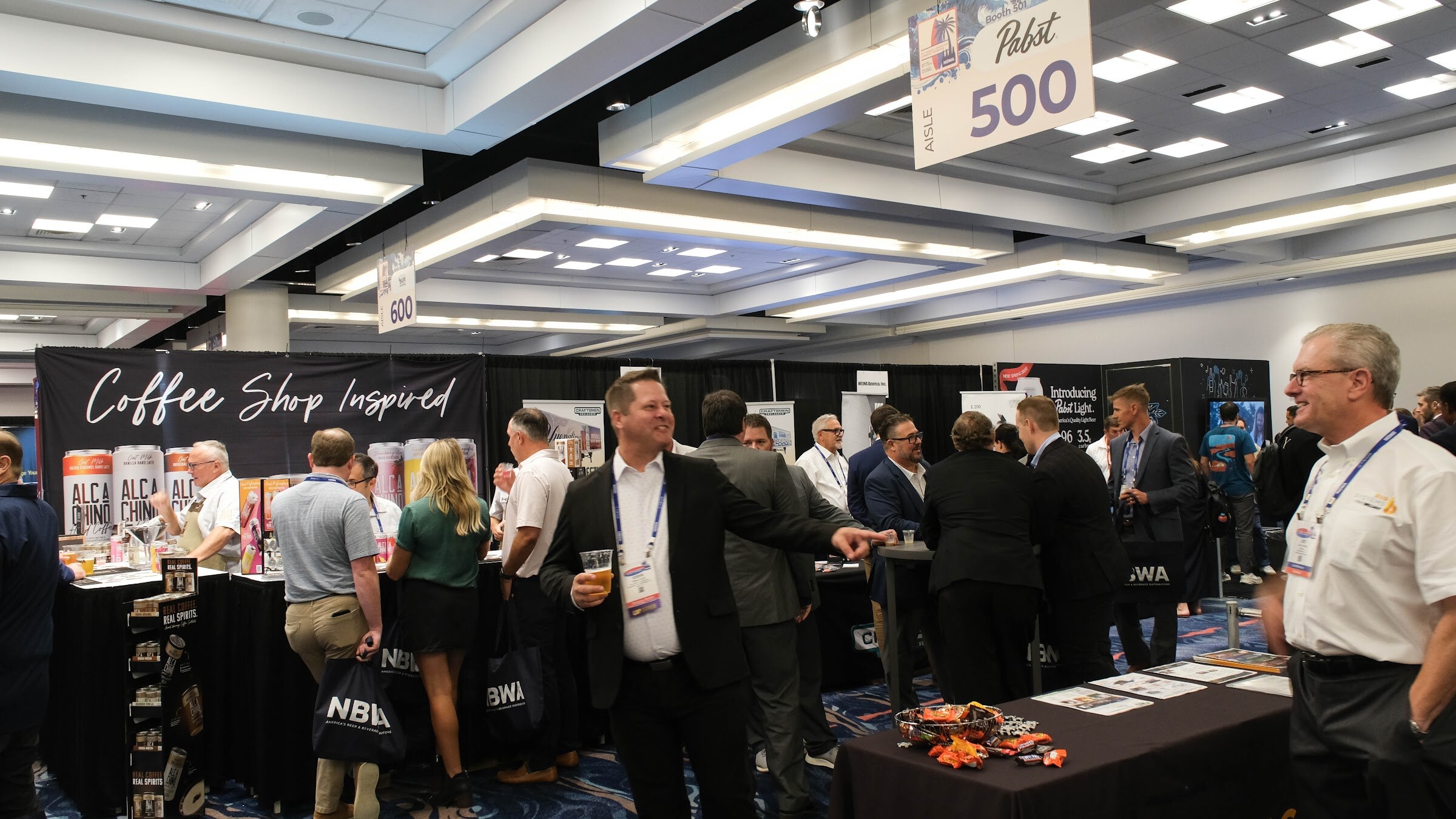 Crowded exhibit hall aisle with attendees holding NBWA tote bags and booths for coffee-inspired beverages in the background.