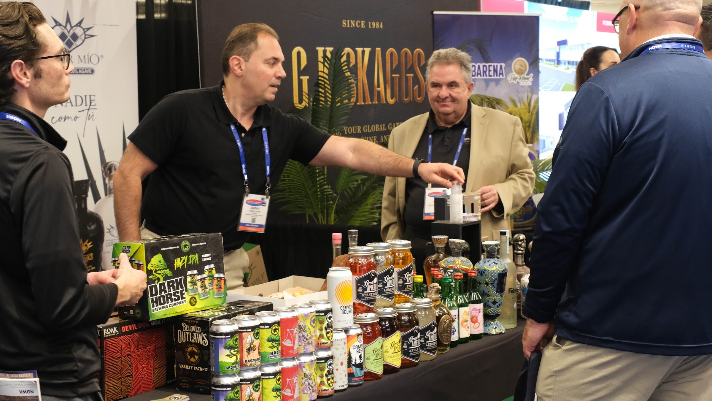 Man in a black polo with a lanyard pointing at products on a table of canned cocktails and tequila bottles, with another man in a tan jacket looking on.