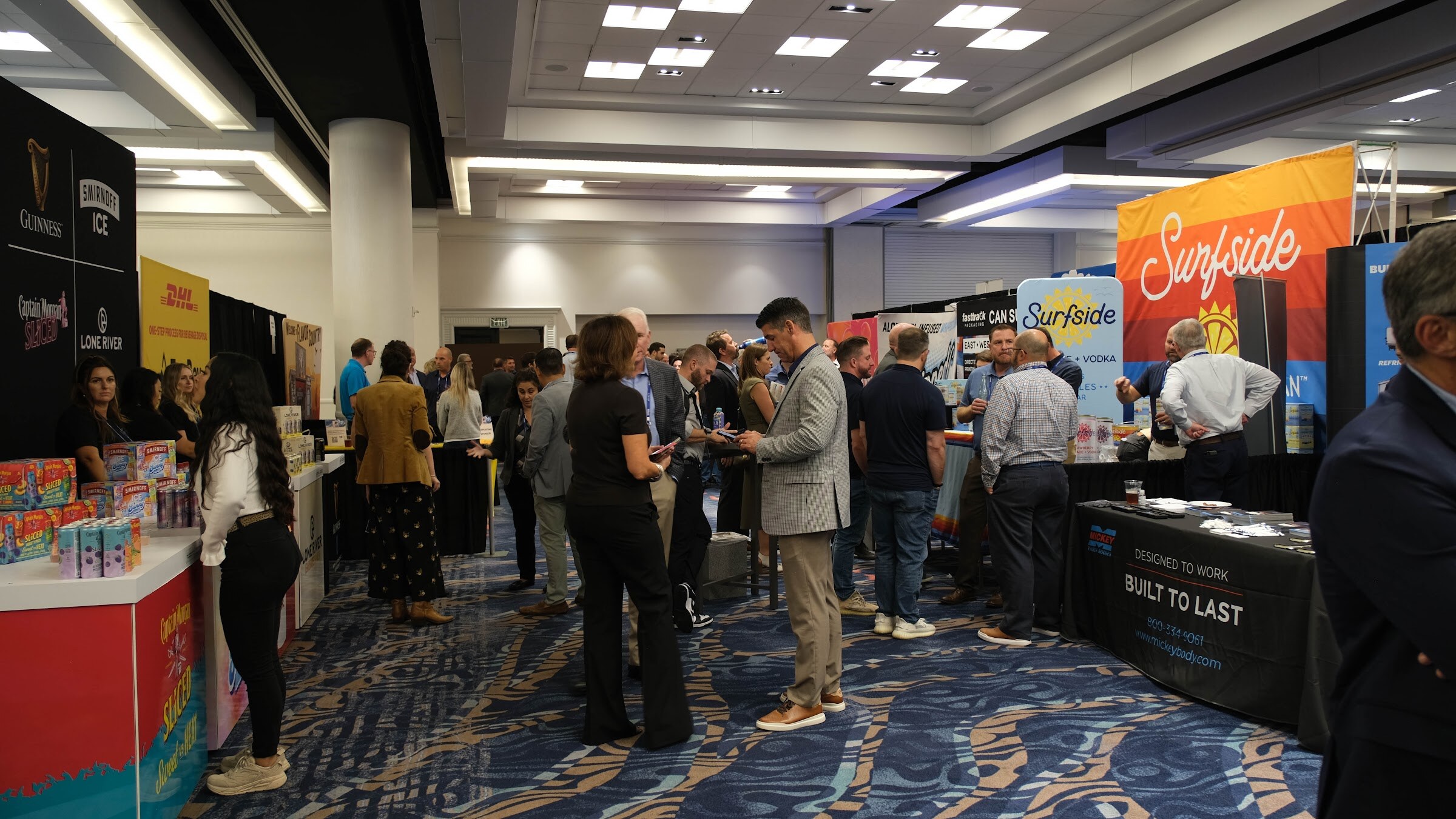 Wide view of an exhibit hall with Surfside, Guinness and other booths and attendees in the aisle on a blue patterned carpet.