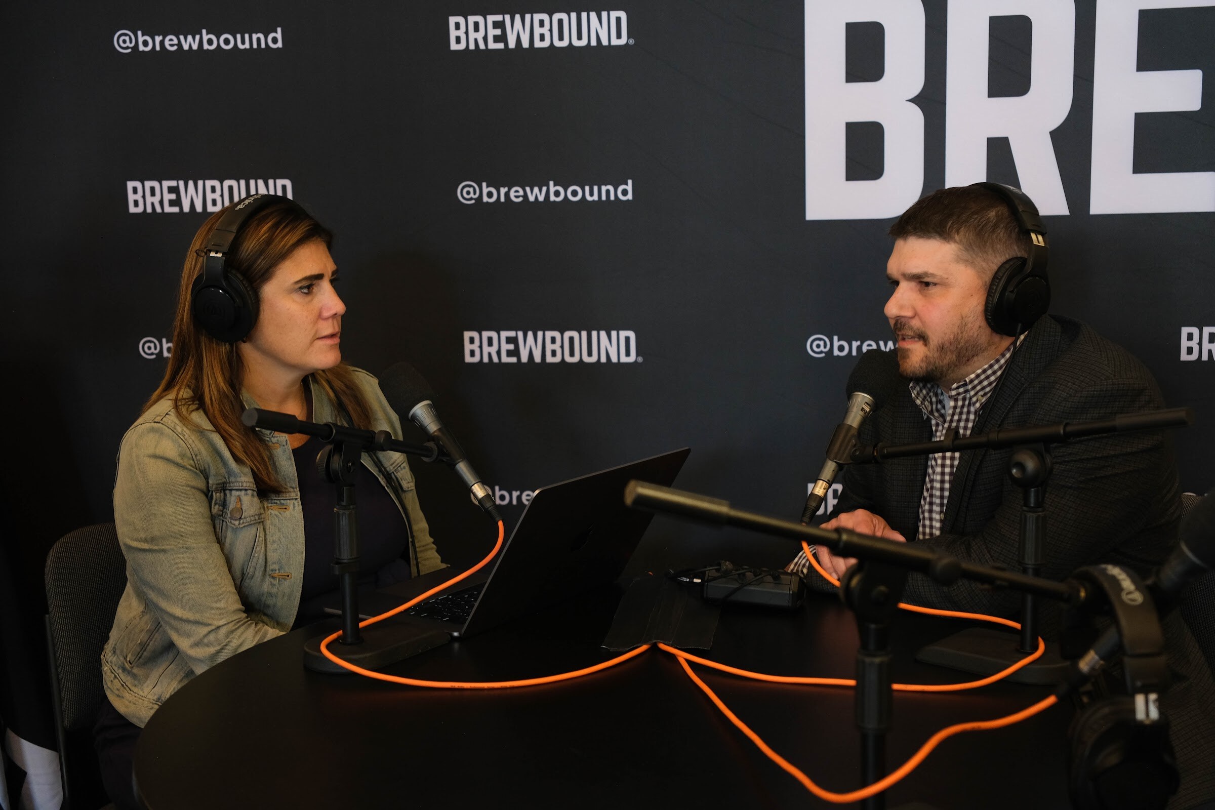 Woman in a denim jacket and a man in a dark jacket wearing headphones at a black table with microphones in front of a Brewbound backdrop.
