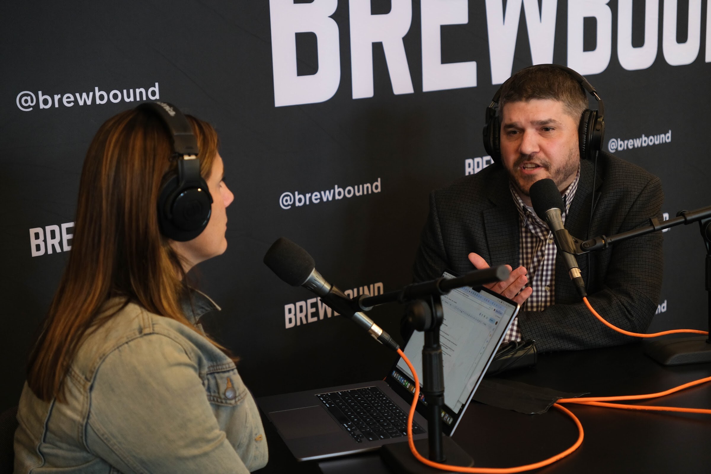 Man with a microphone and headphones speaking, with the host in the foreground, in front of a Brewbound backdrop.