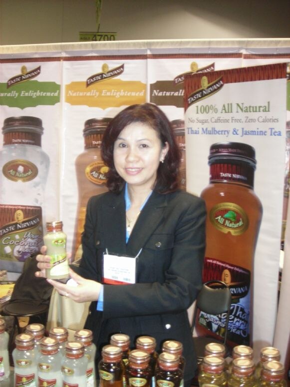 A smiling woman in a dark blazer holds a bottle of Taste Nirvana coconut drink. Taste Nirvana banners and rows of bottles surround her.