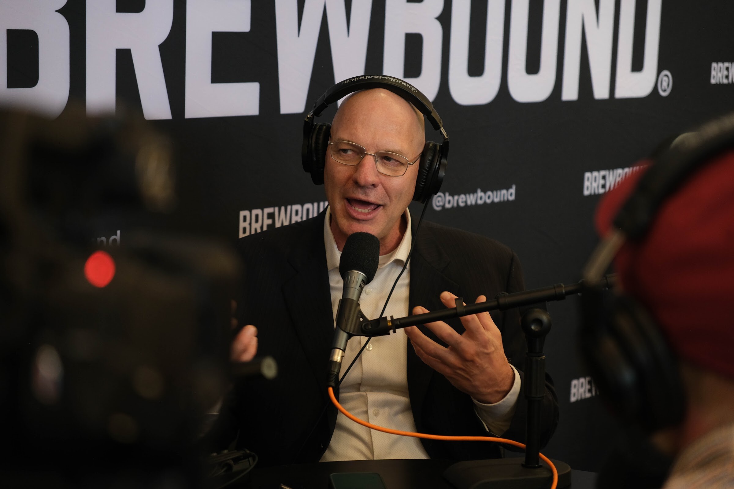 Gray-haired man with glasses in a dark jacket and headphones speaking into a microphone in front of a Brewbound backdrop.