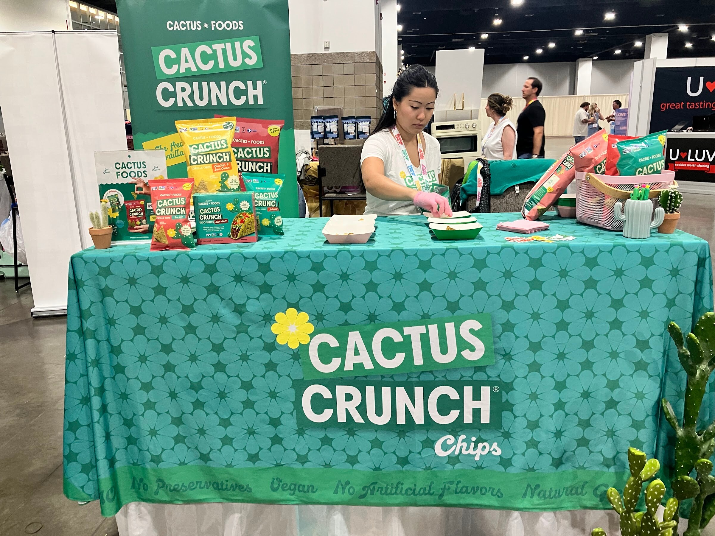 Woman prepares samples at a teal Cactus Crunch table with bags of chips and a Cactus Foods banner.