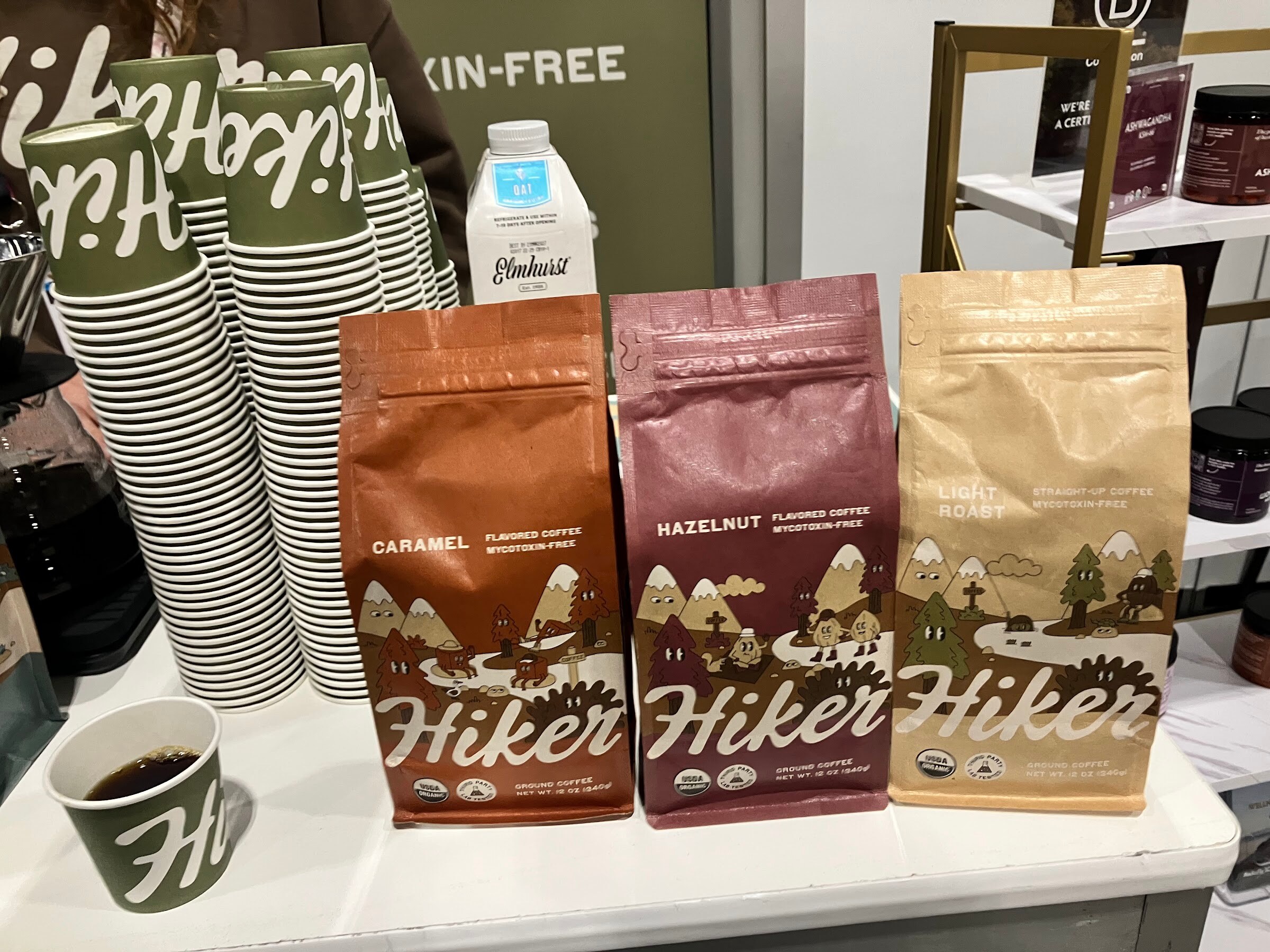 Three bags of Hiker coffee beside stacks of green Hiker cups and a sample of brewed coffee.