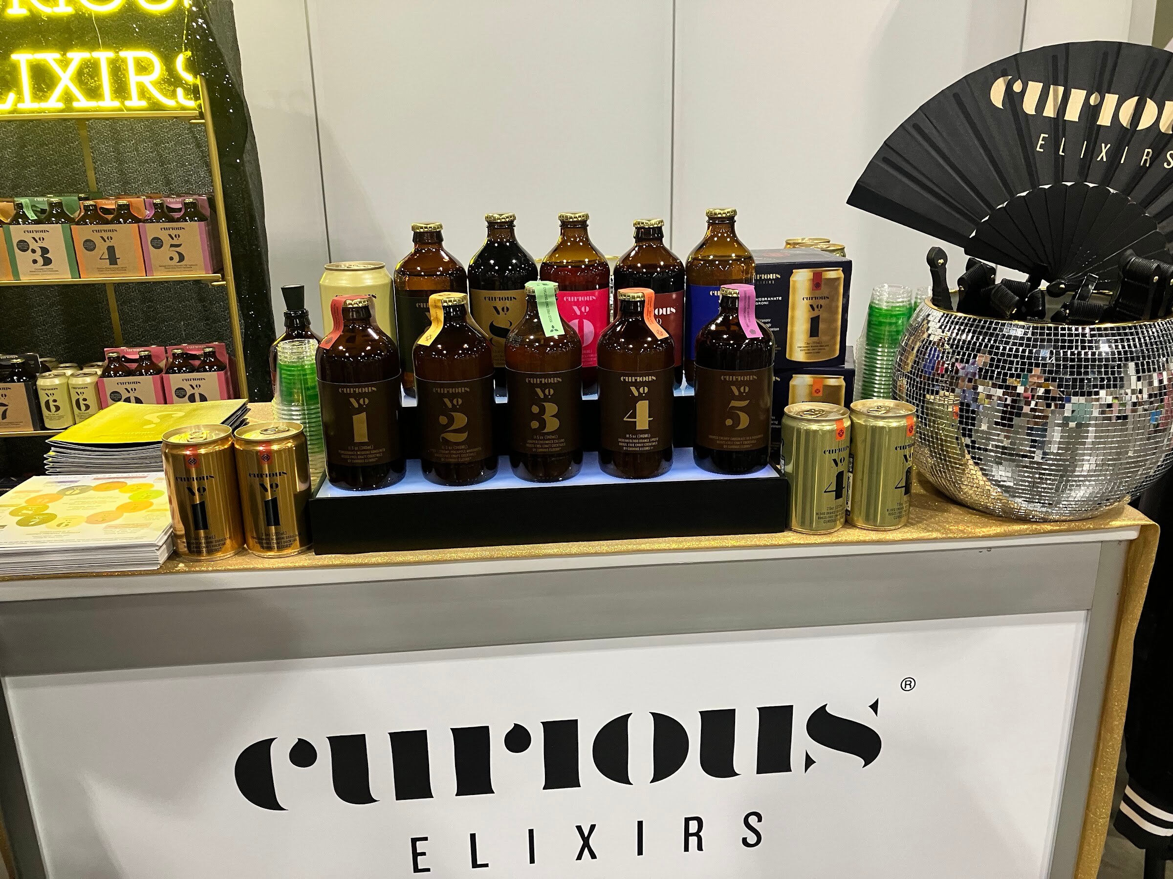 Rows of amber Curious Elixirs bottles and gold cans on a counter with a disco ball and a black fan.