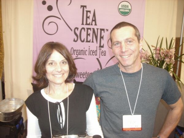 A smiling woman and man stand together in front of a pink Tea Scene Organic Iced Tea banner with a USDA Organic seal.