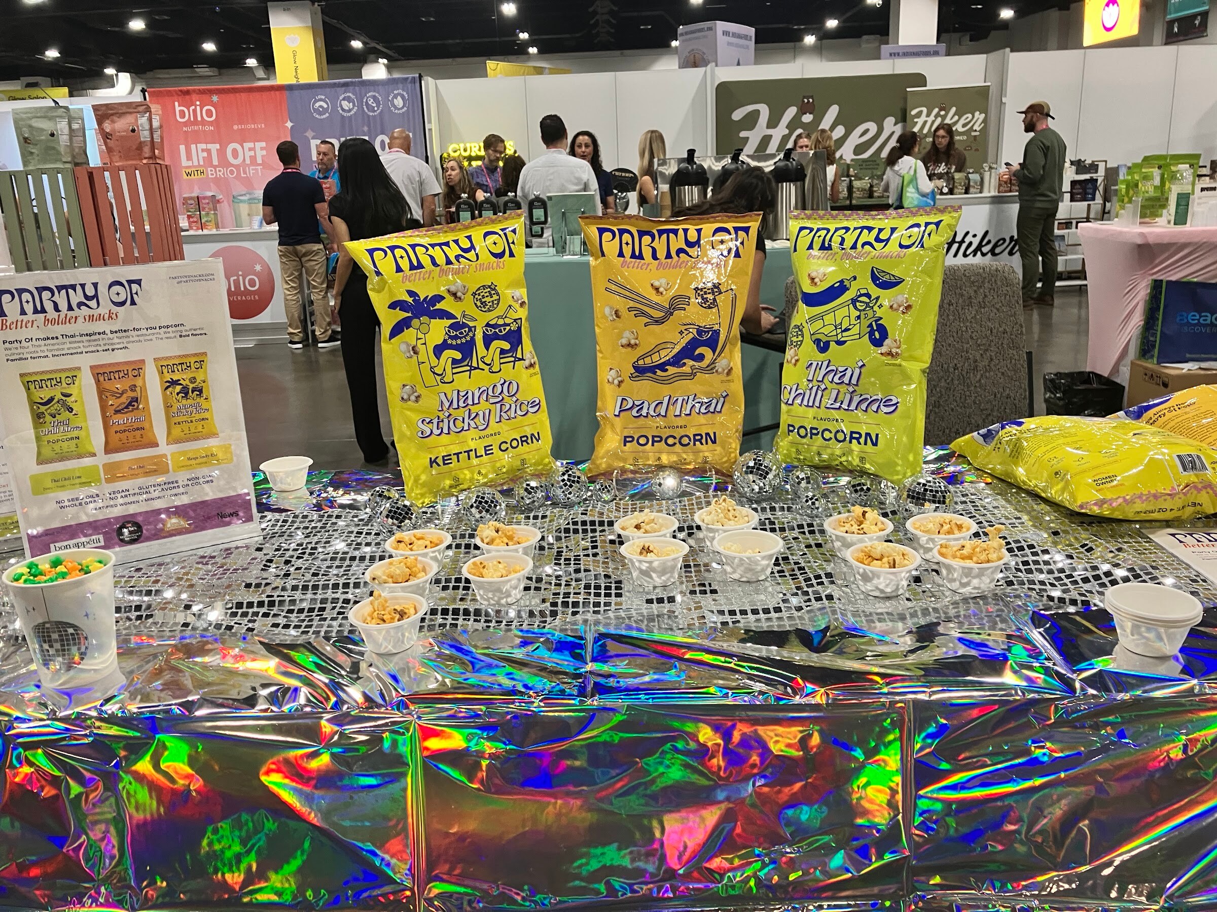 Yellow bags of Party Of popcorn and sample cups on a holographic tablecloth on the busy show floor.