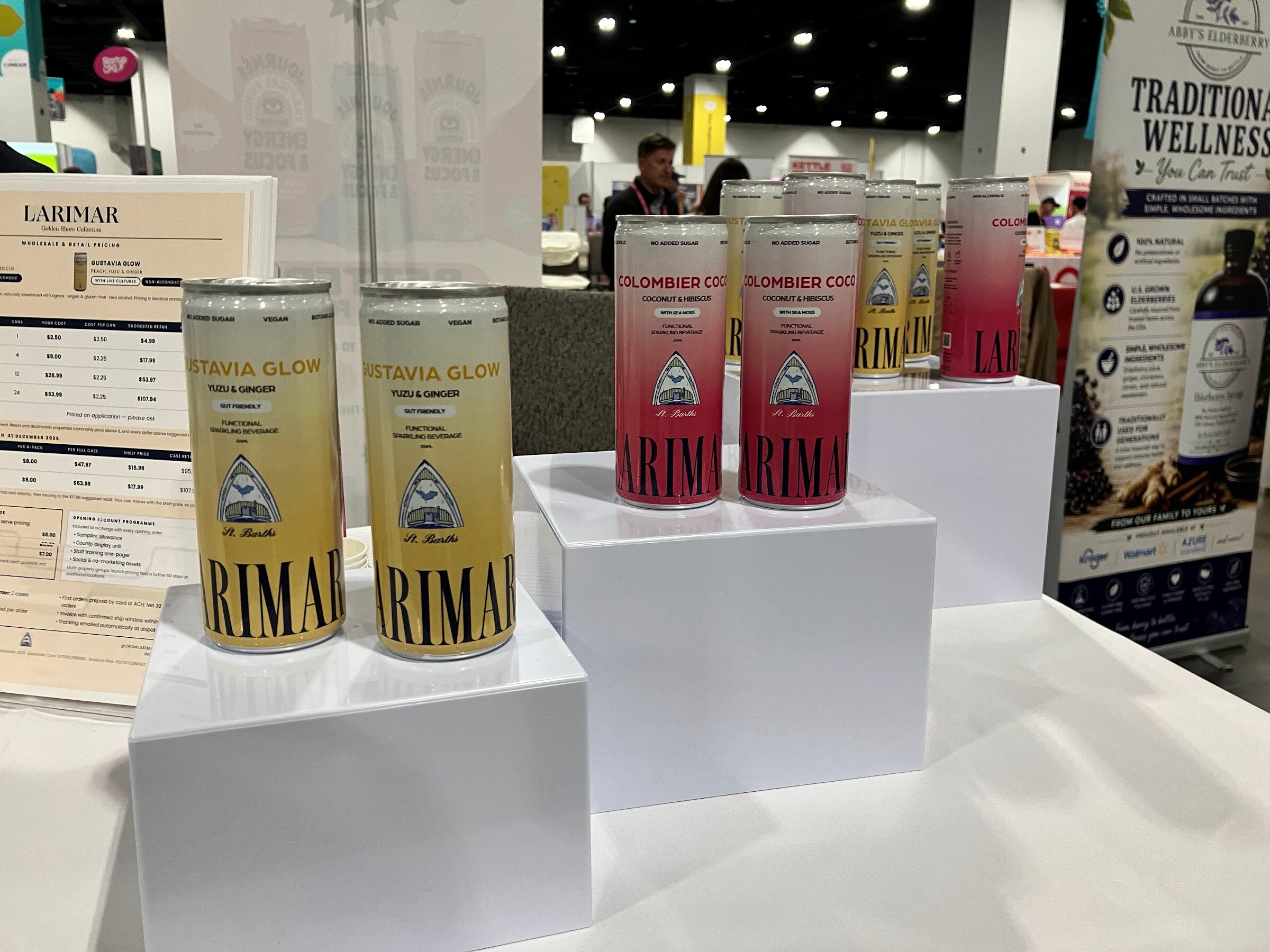 Cans of Larimar in yellow and pink flavors on white display risers with a price sheet beside them.