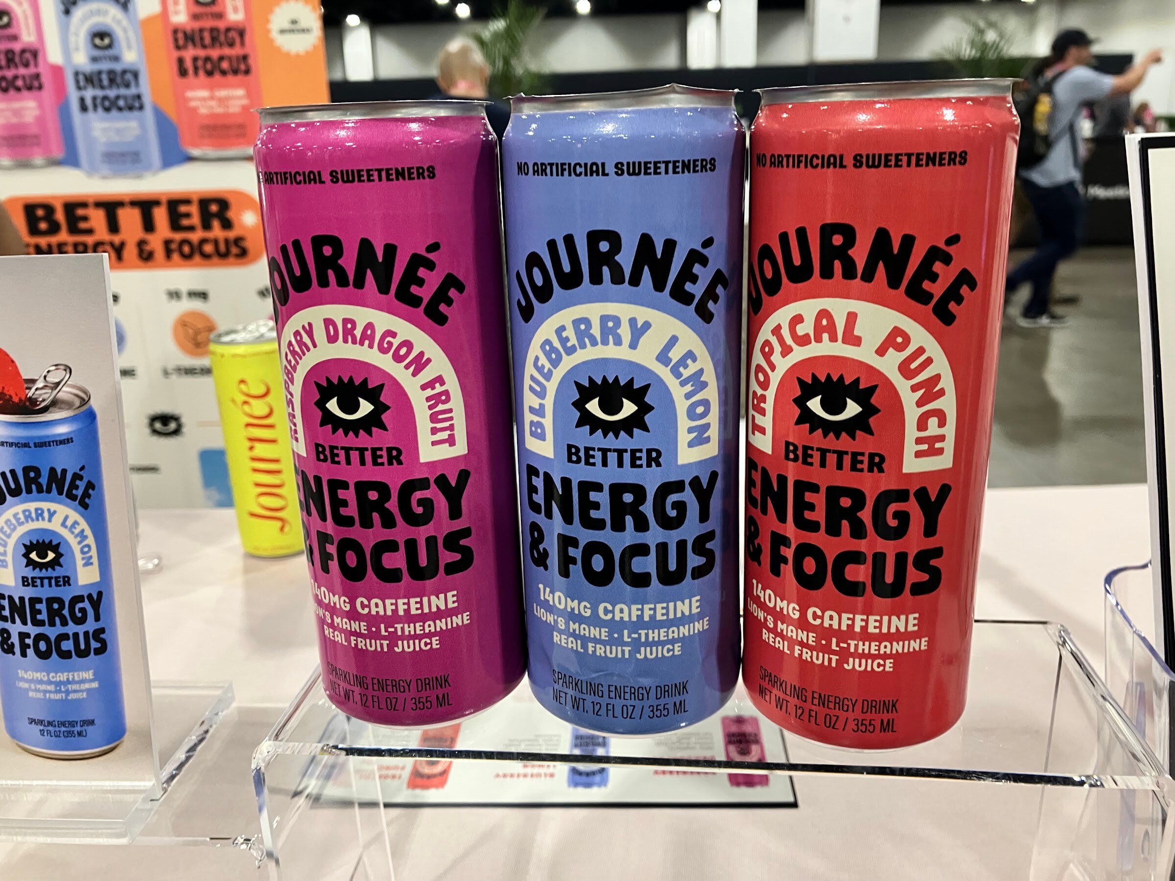 Three cans of Journée energy drink in dragon fruit, blueberry lemon and tropical punch on an acrylic stand.