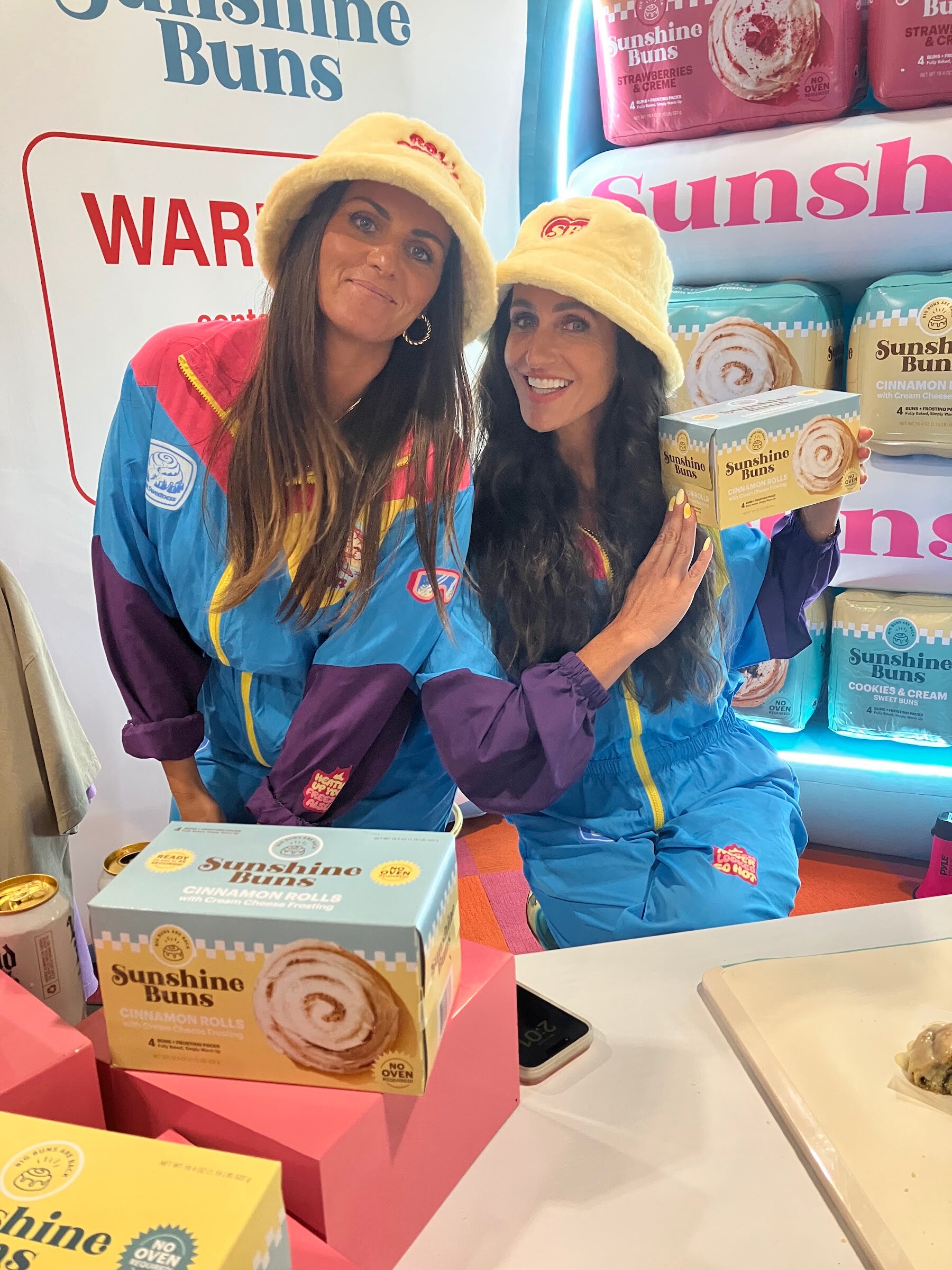 Two smiling women in yellow bucket hats and colorful track jackets pose with boxes of Sunshine Buns cinnamon rolls.