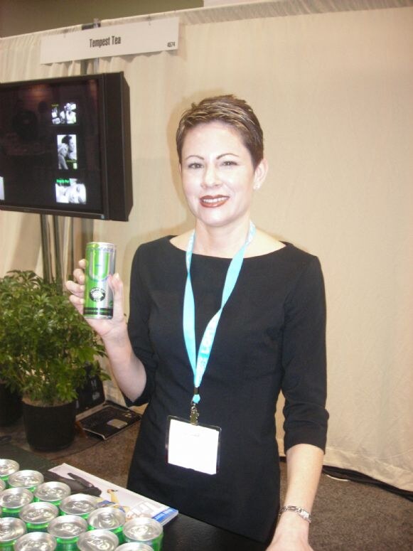 A woman in a black dress and lanyard holds a green can at the Tempest Tea booth. Rows of green cans sit on the table and a monitor and plant are behind her.