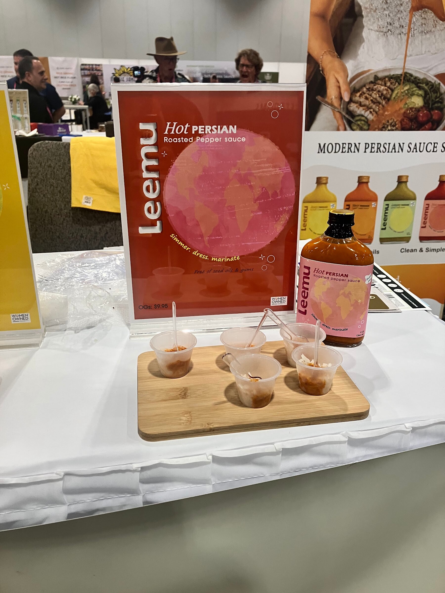 Sample cups on a wooden board in front of a Leemu Hot Persian sign and a bottle of sauce.