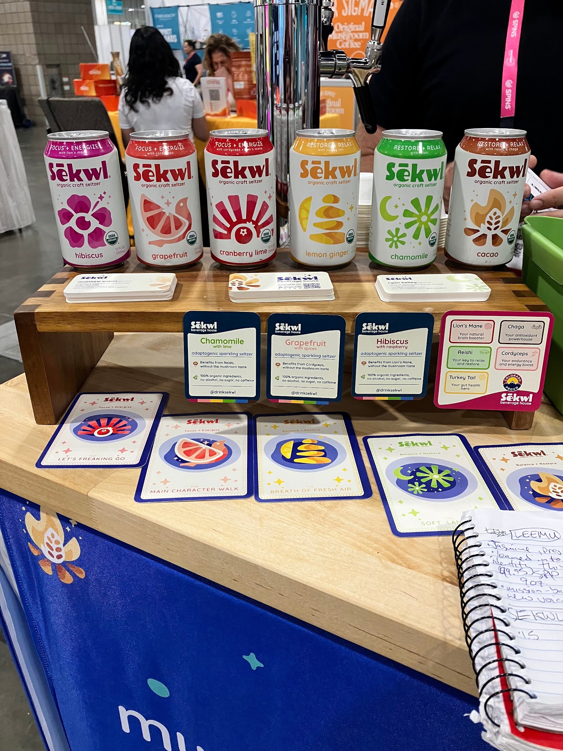 Six cans of Sekwel soda in hibiscus, grapefruit and other flavors on a wooden riser above flavor cards.