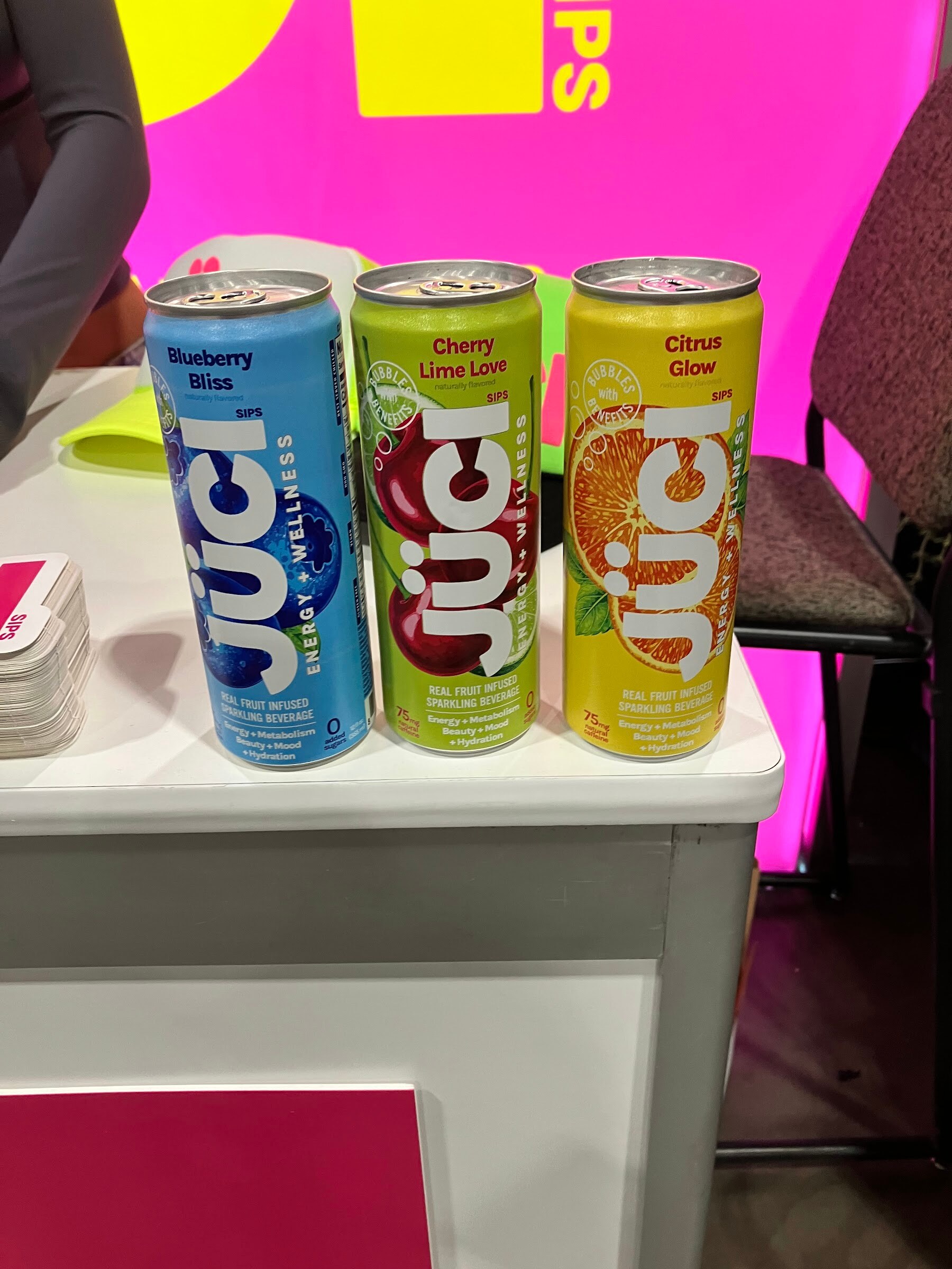 Three cans of Juci in blue, green and yellow on a white table against a pink backdrop.