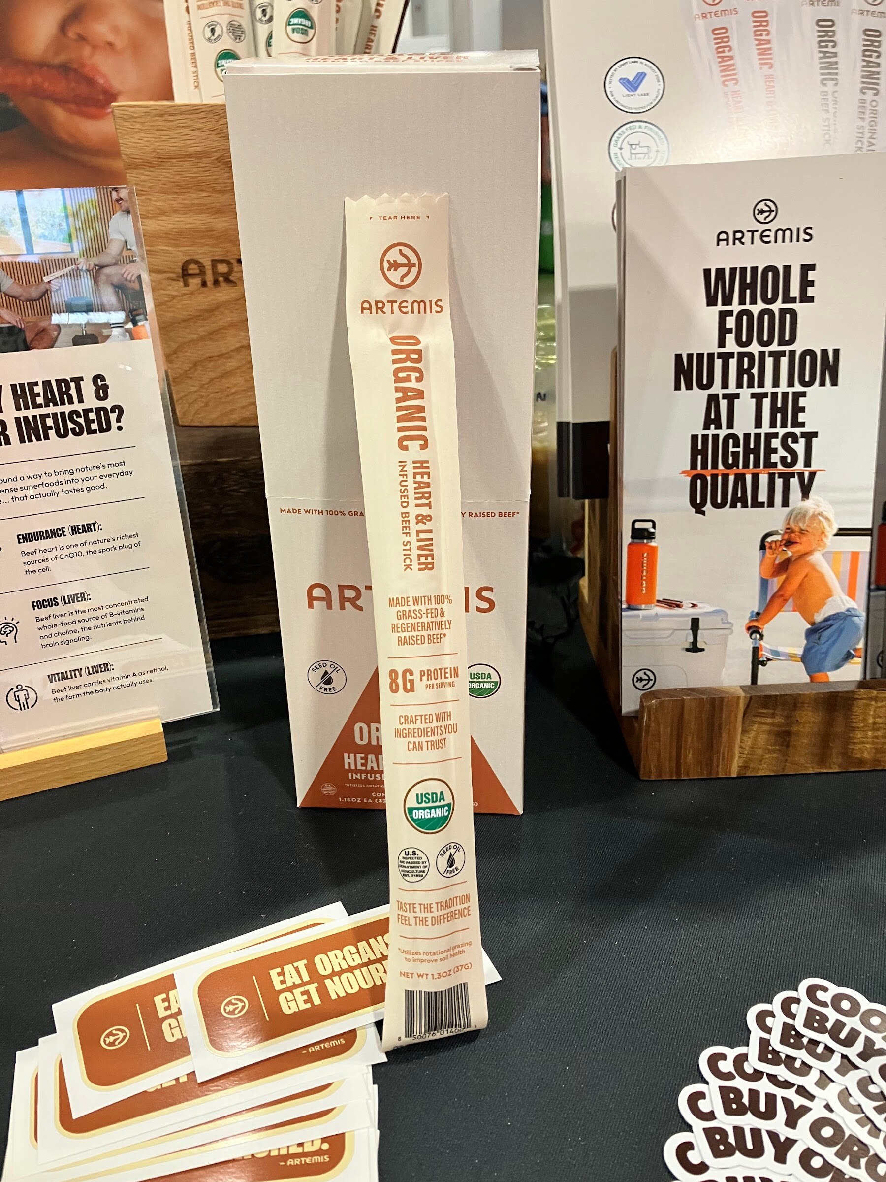 An Artemis organic beef stick standing in front of boxes and a sign reading Whole food nutrition at the highest quality.